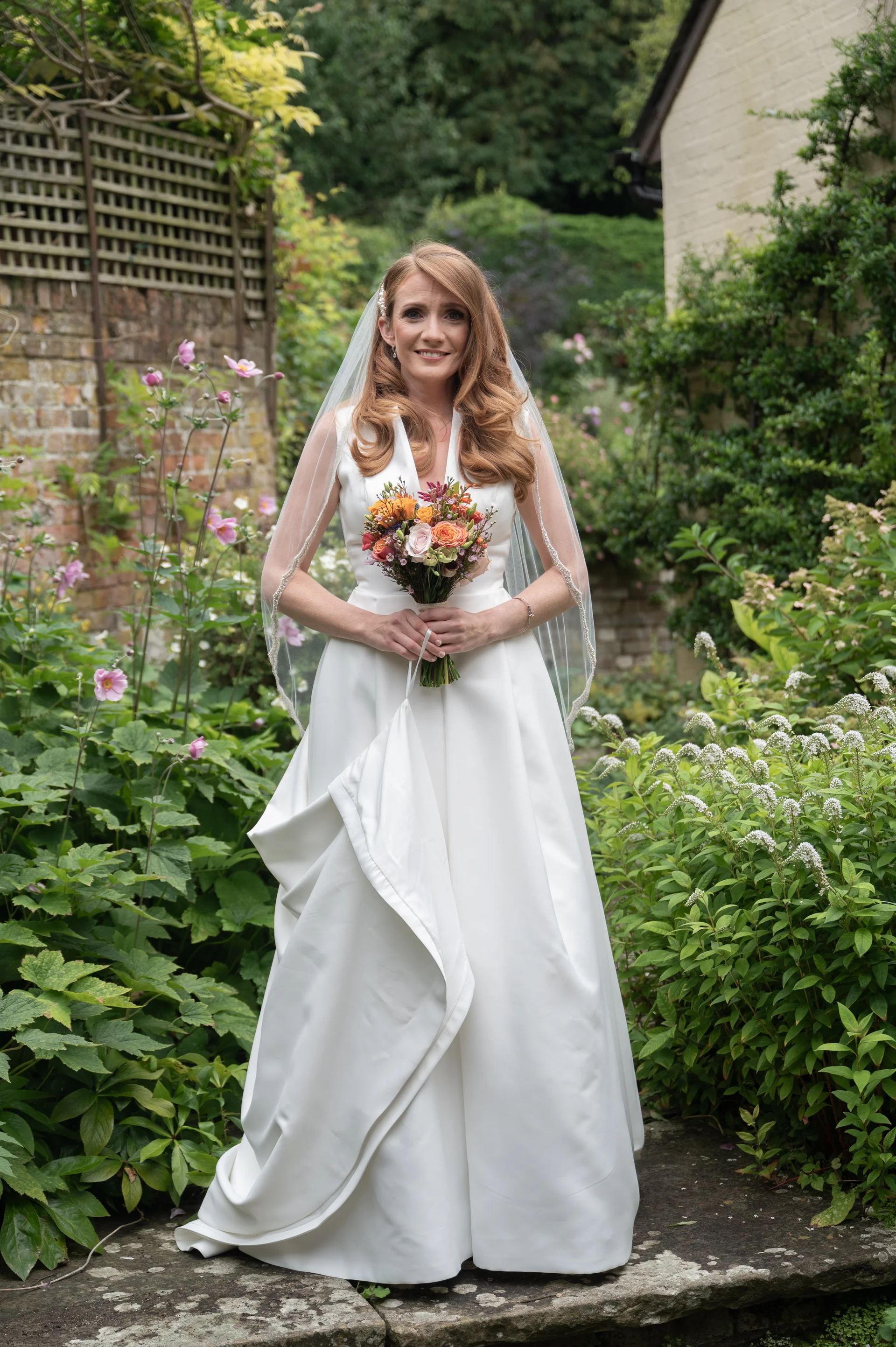 A smiling bride stands in a lush garden holding a vibrant bouquet, wearing a satin V-neck gown and a delicate veil.
