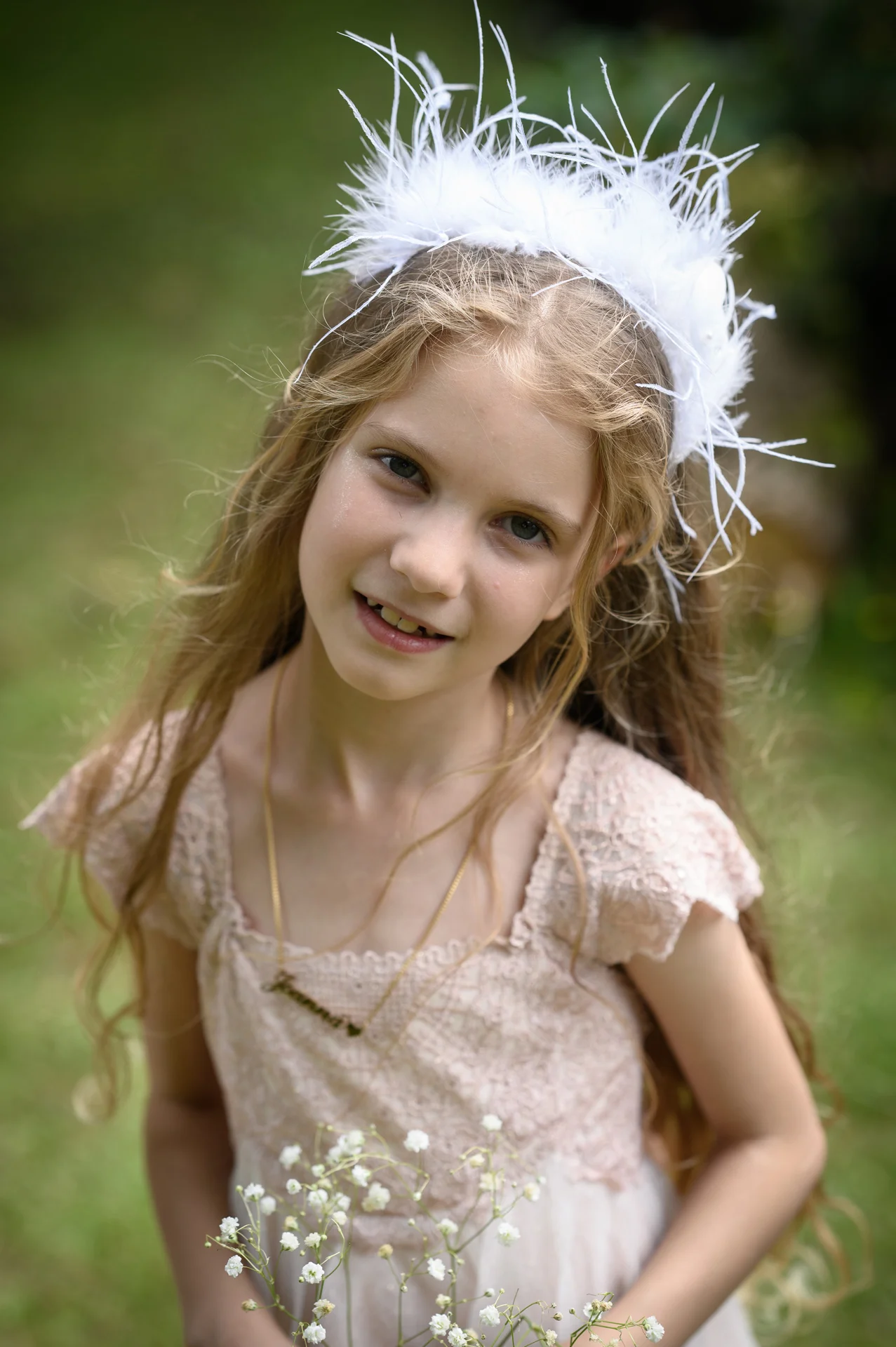 A young flower girl with long blonde hair smiles gently while wearing a white feather headpiece and holding delicate baby's breath flowers outdoors.