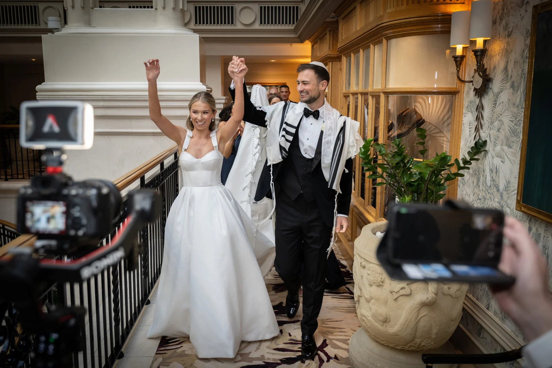 A newlywed couple joyfully walks hand in hand while holding their arms up, captured on a balcony inside a luxury hotel venue.