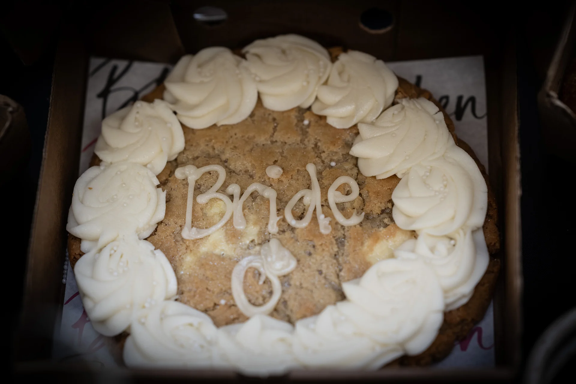 A close-up overhead shot of a giant cookie cake decorated with white piped frosting swirls and the word bride written in icing.