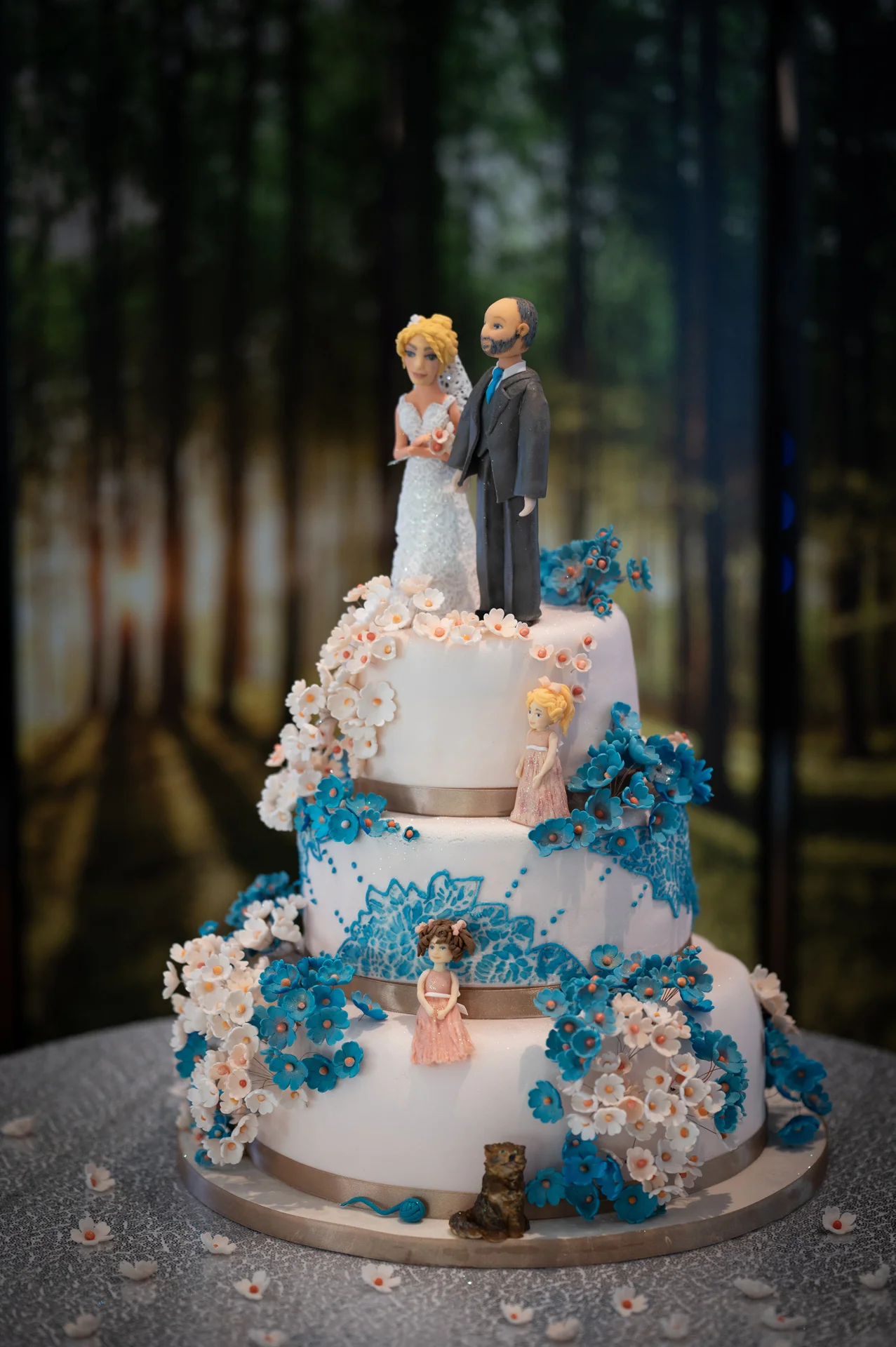 A detailed three-tier wedding cake decorated with blue and white sugar flowers features custom bride and groom figurines on top.