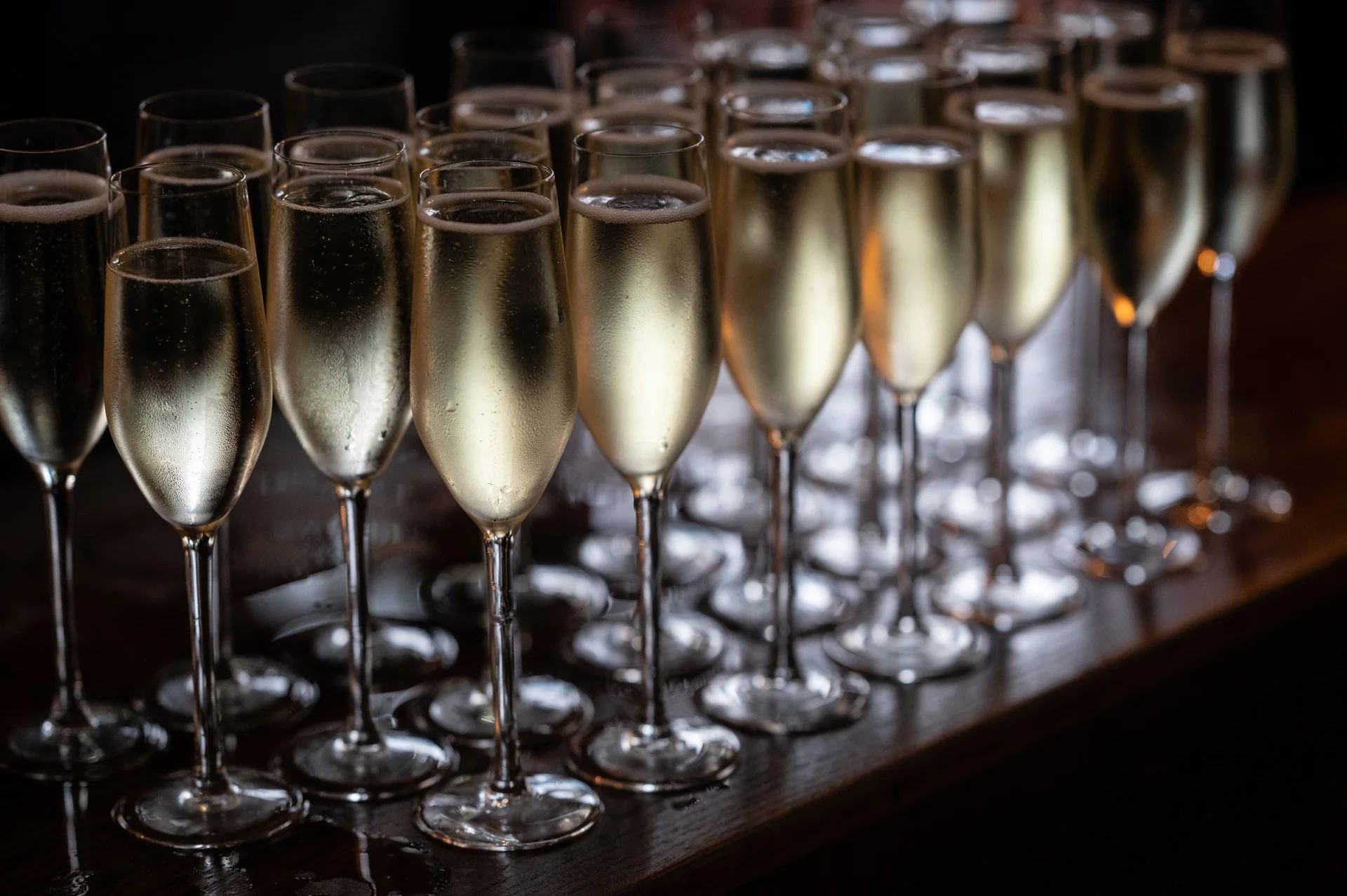 An elegant arrangement of crystal champagne flutes filled with sparkling wine lines a wooden bar under moody lighting.