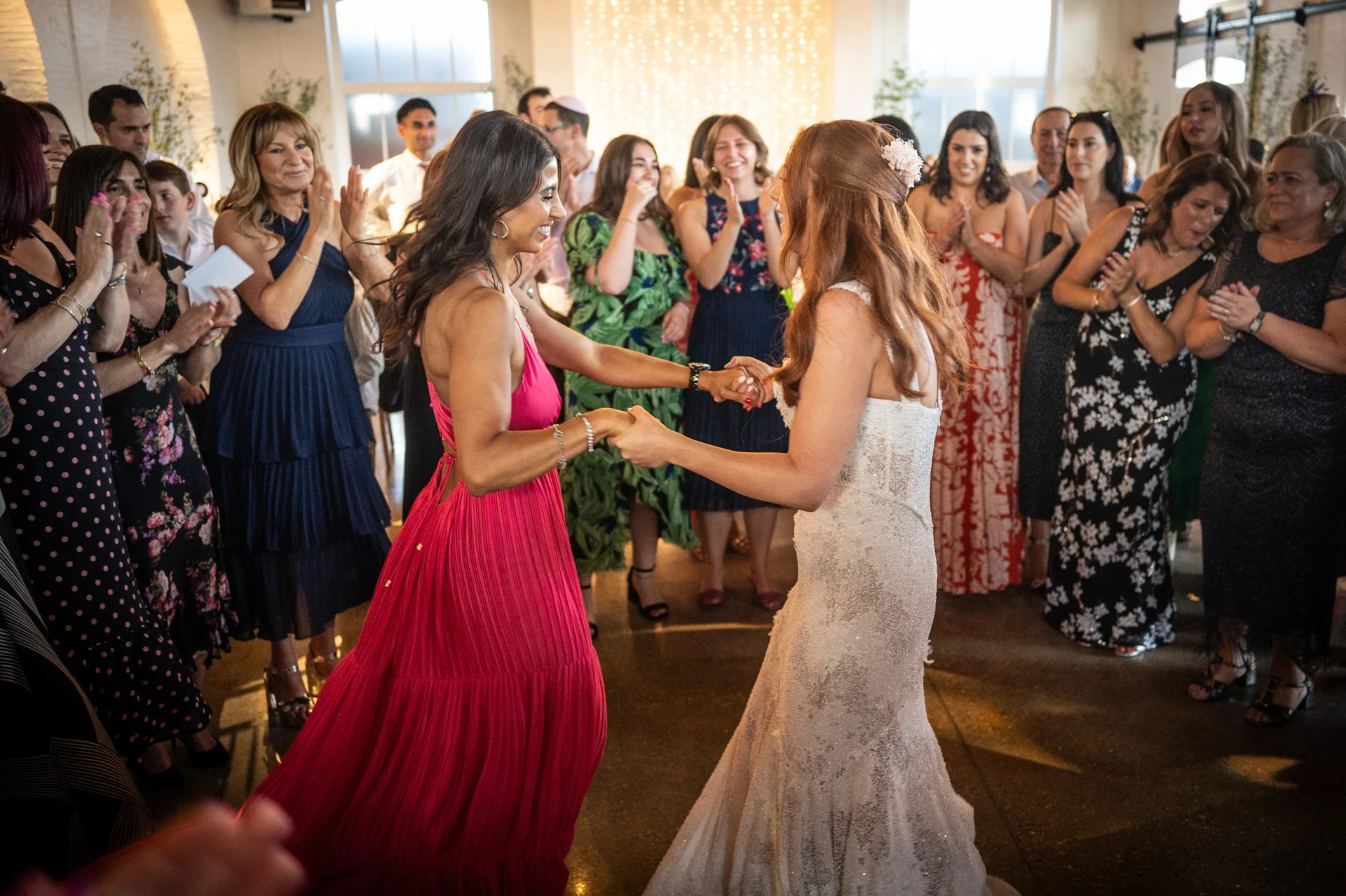 Two brides joyfully hold hands and dance together while wedding guests clap and cheer in an industrial venue.