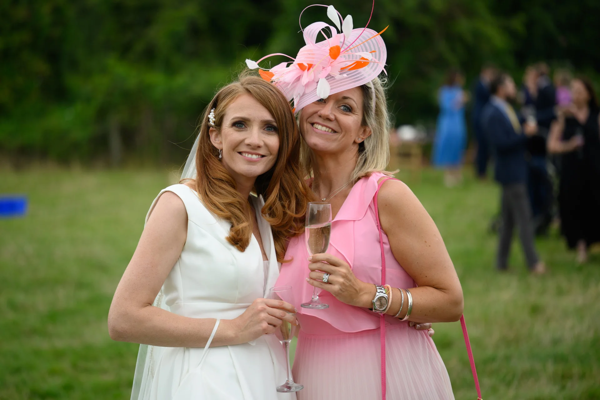 A smiling bride in a white v-neck crepe gown and a guest wearing a pink dress with a dramatic feathered fascinator pose together outdoors while holding champagne flutes during a drinks reception.