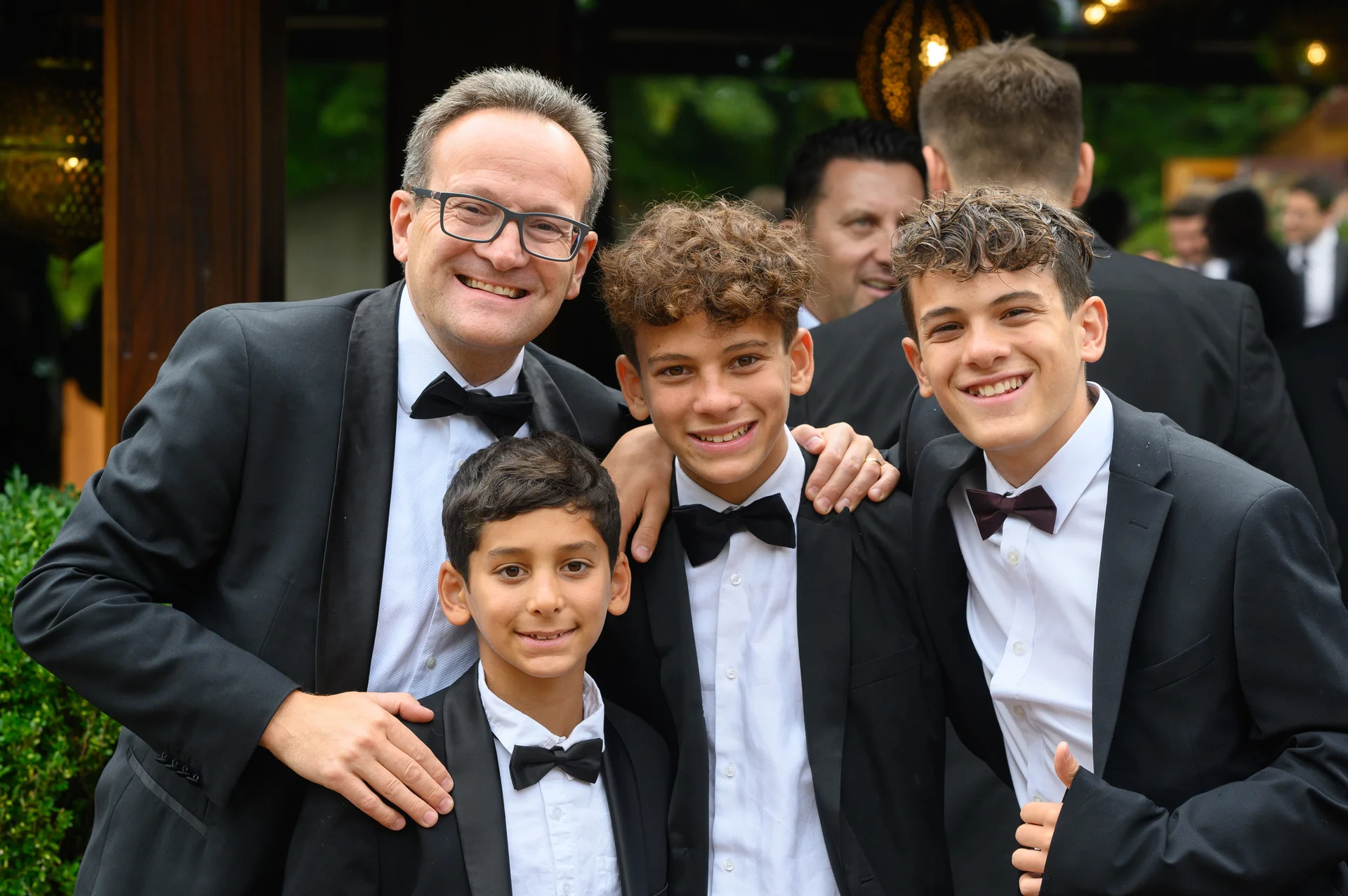 An adult man and three smiling boys dressed in smart black tuxedos with bow ties pose together at a wedding celebration.