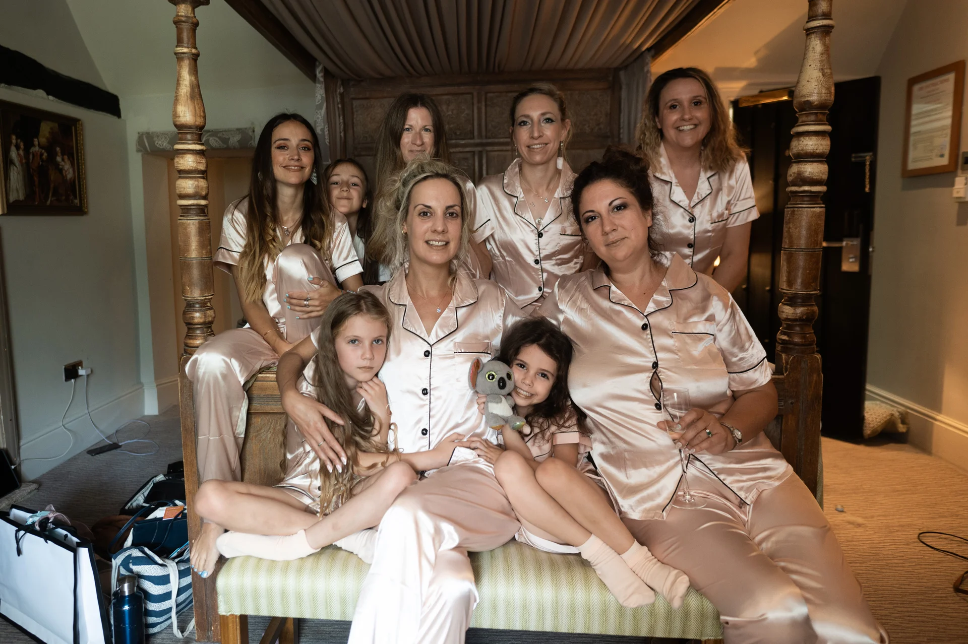 A bridal party and young flower girls pose together on a wooden bench in matching pink satin pyjamas during morning preparations.