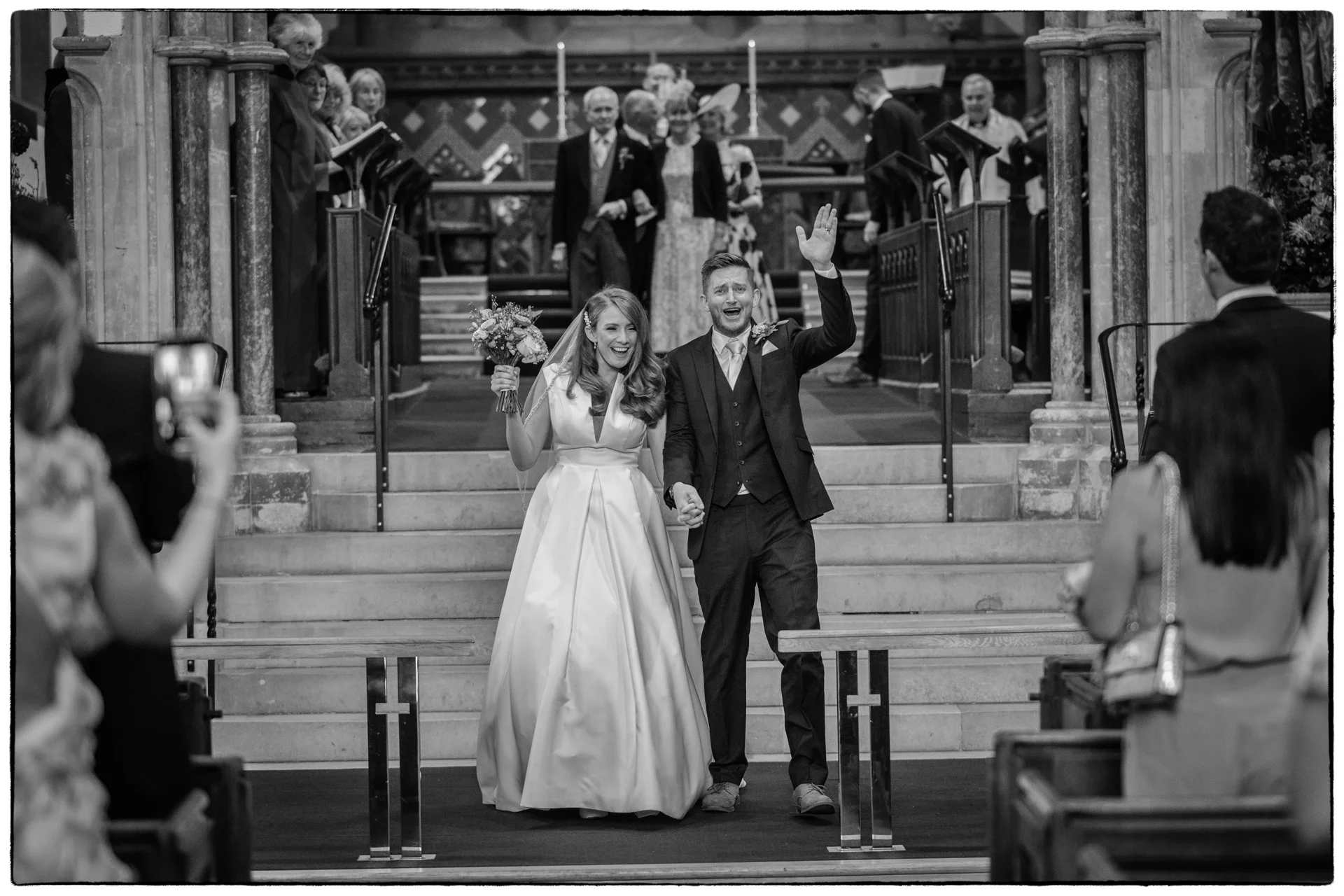A joyful bride and groom walk hand in hand down the aisle of a historic church interior, laughing and waving to cheering guests.