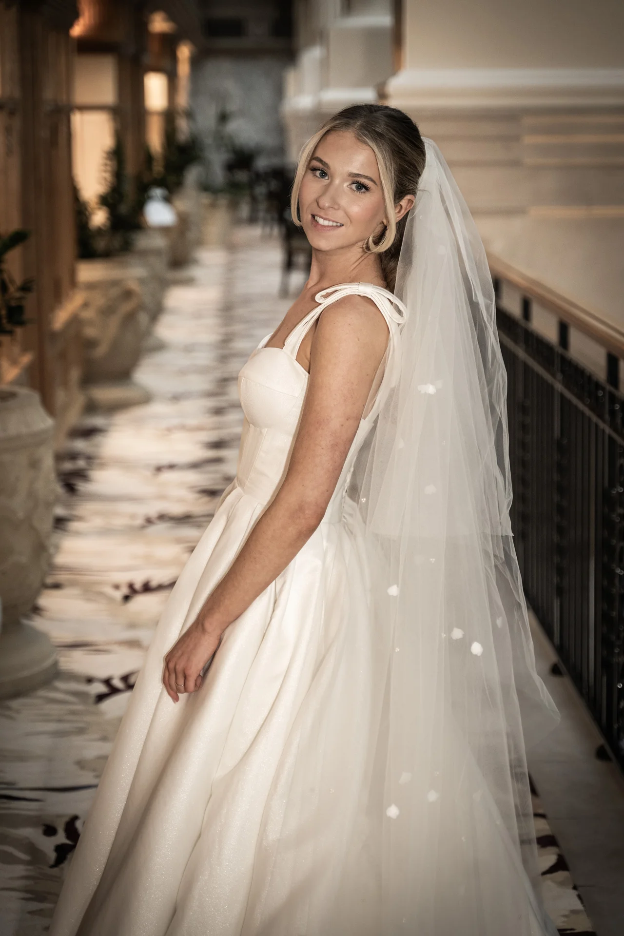A smiling bride wearing an a-line gown and a dotted veil stands in a grand hallway, looking back over her shoulder towards the camera.