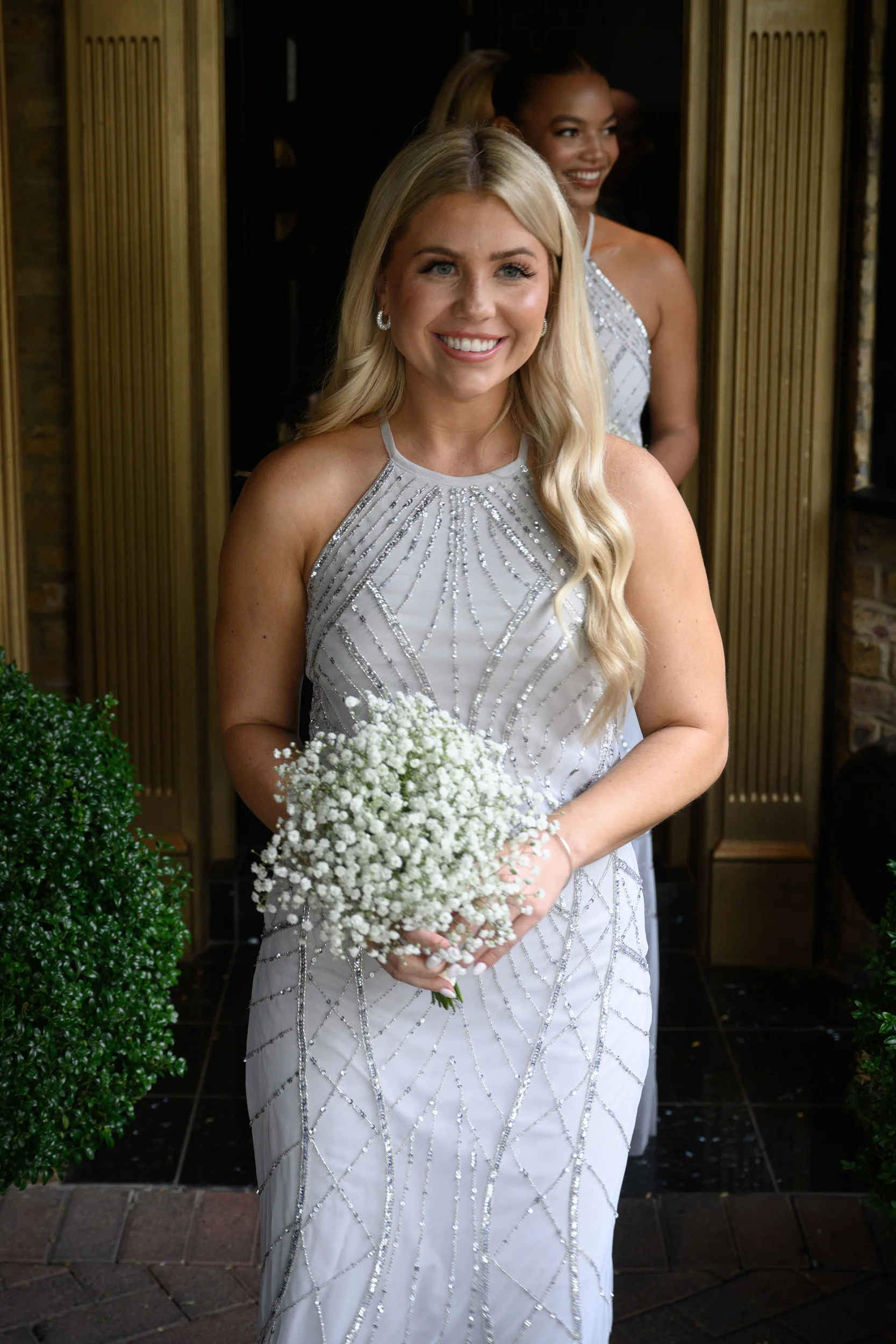 A smiling blonde bridesmaid holding a gypsophila posy bouquet wears a halter-neck beaded silver dress as she steps through a doorway.