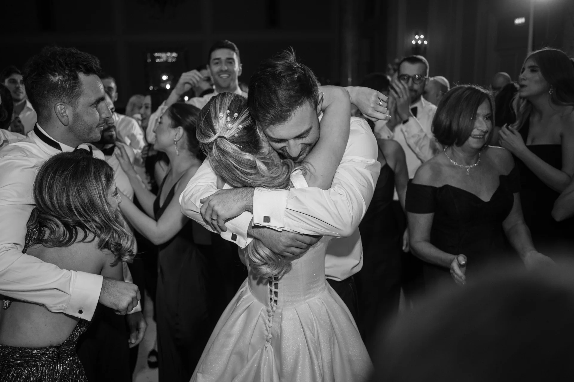A newlywed couple share a tight, joyful embrace in the middle of a lively evening reception surrounded by celebrating wedding guests in monochrome.