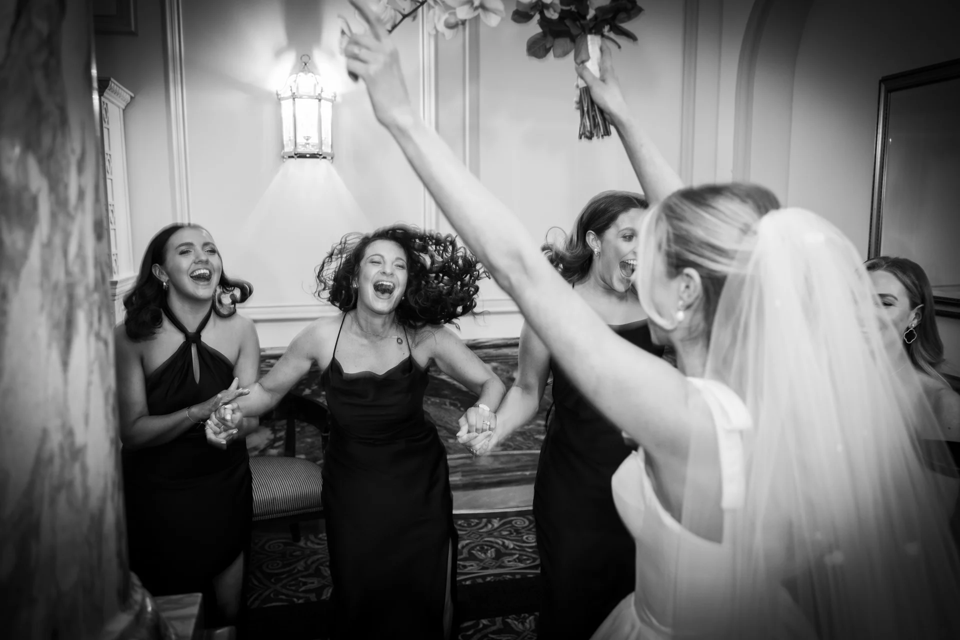 A joyous bride in a wedding gown with a veil celebrates and holds up her bouquet while surrounded by laughing bridesmaids in elegant dresses.