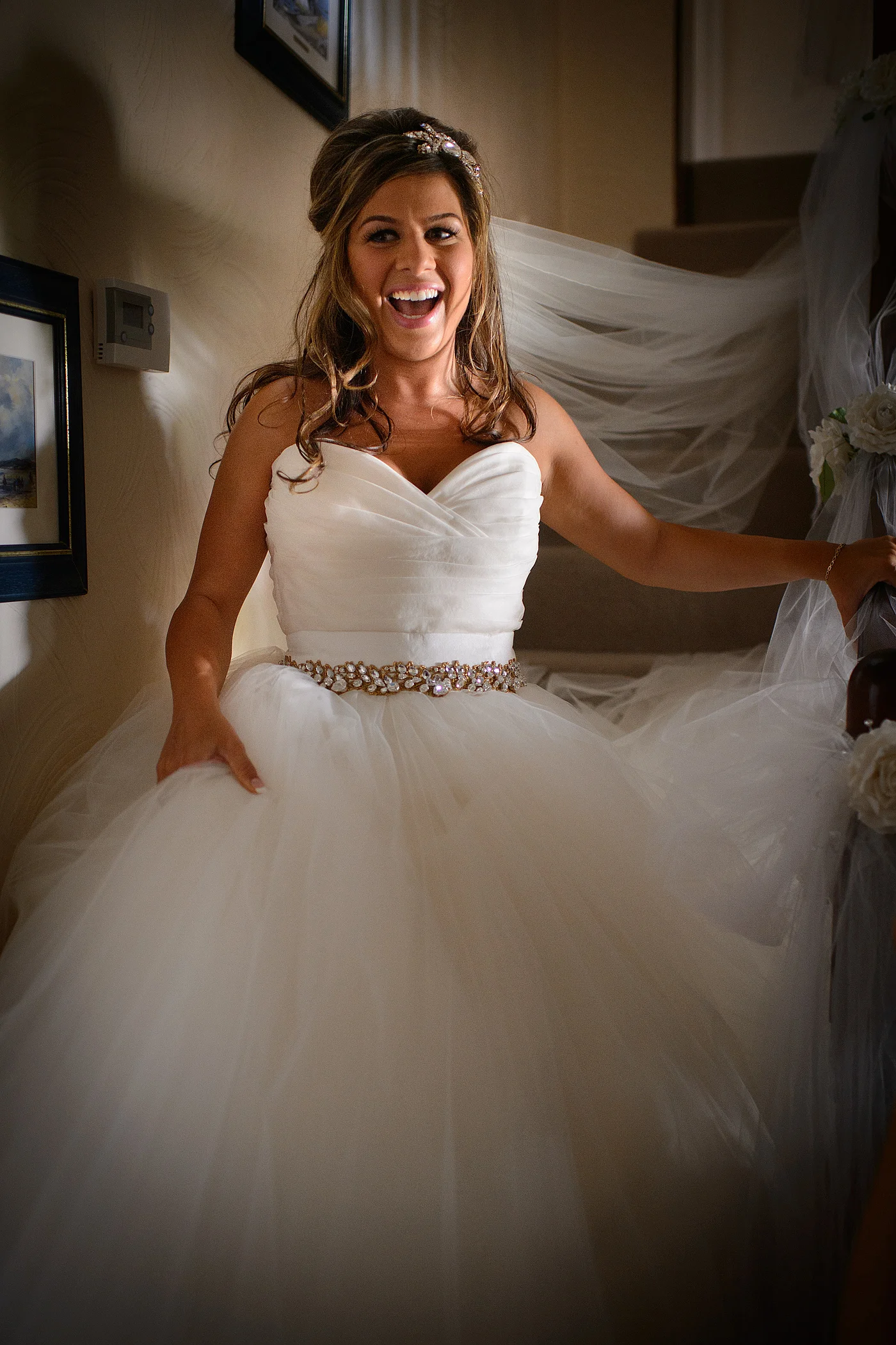 A joyful bride wearing a voluminous ball gown with a crystal belt laughs excitedly while descending a staircase during wedding preparations.