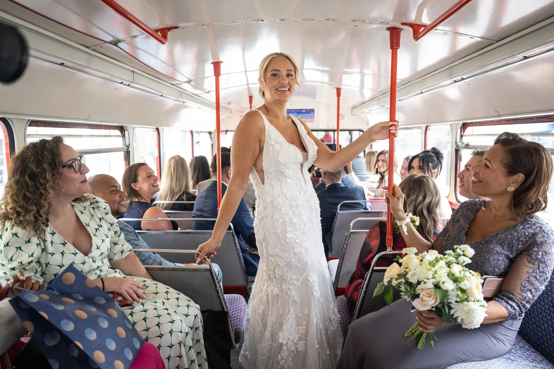 A smiling bride wearing a 3D floral lace wedding dress stands inside a vintage red double decker bus while holding onto a pole, surrounded by laughing guests.