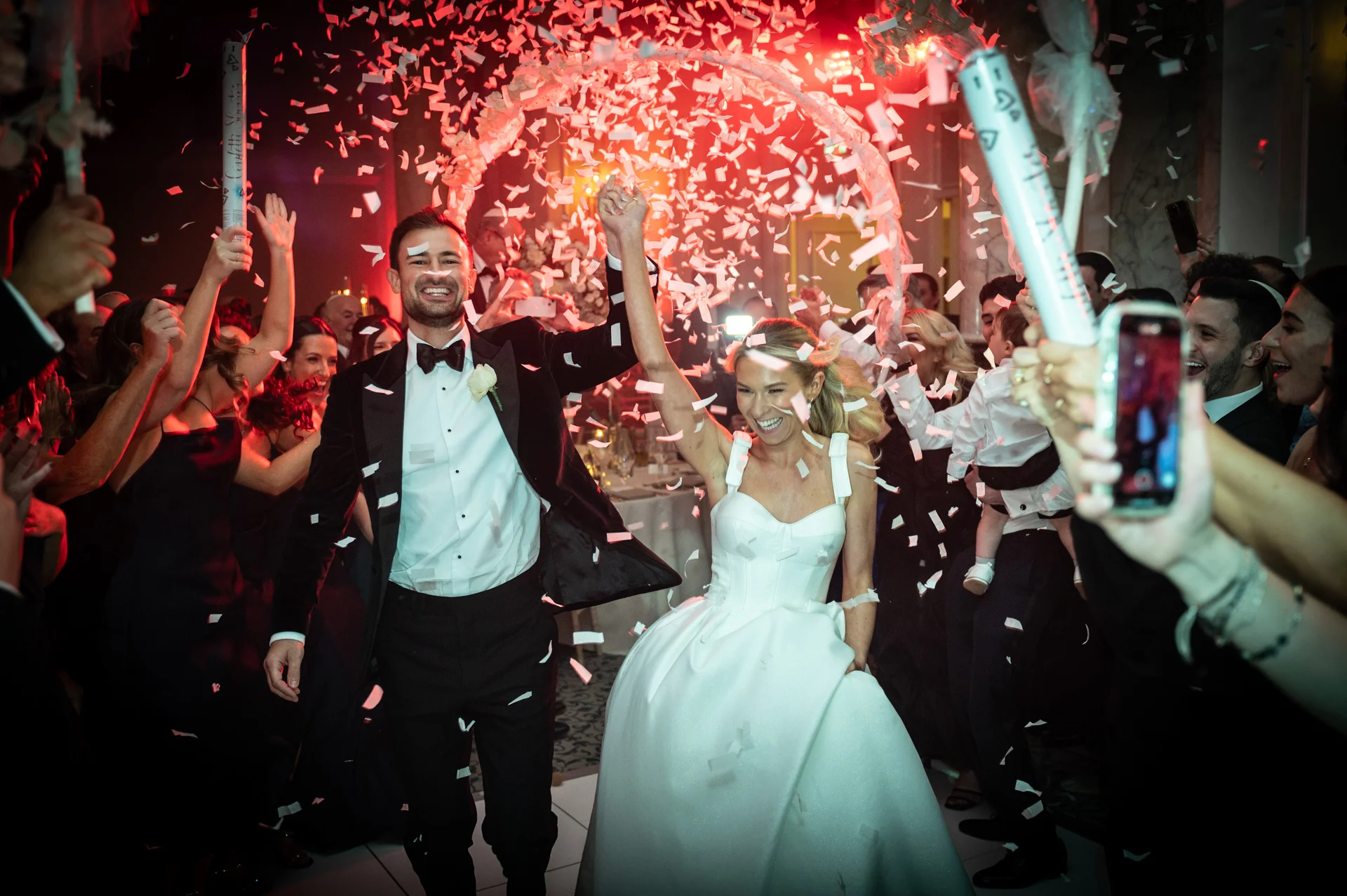 A newlywed couple joyful and laughing as they enter their evening reception amidst a shower of white confetti and cheering guests.