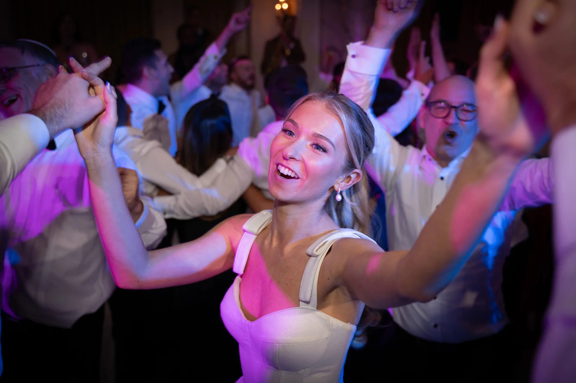 A joyful bride dances with her hands raised on a lively, vibrant dance floor surrounded by cheering guests under colourful lighting.