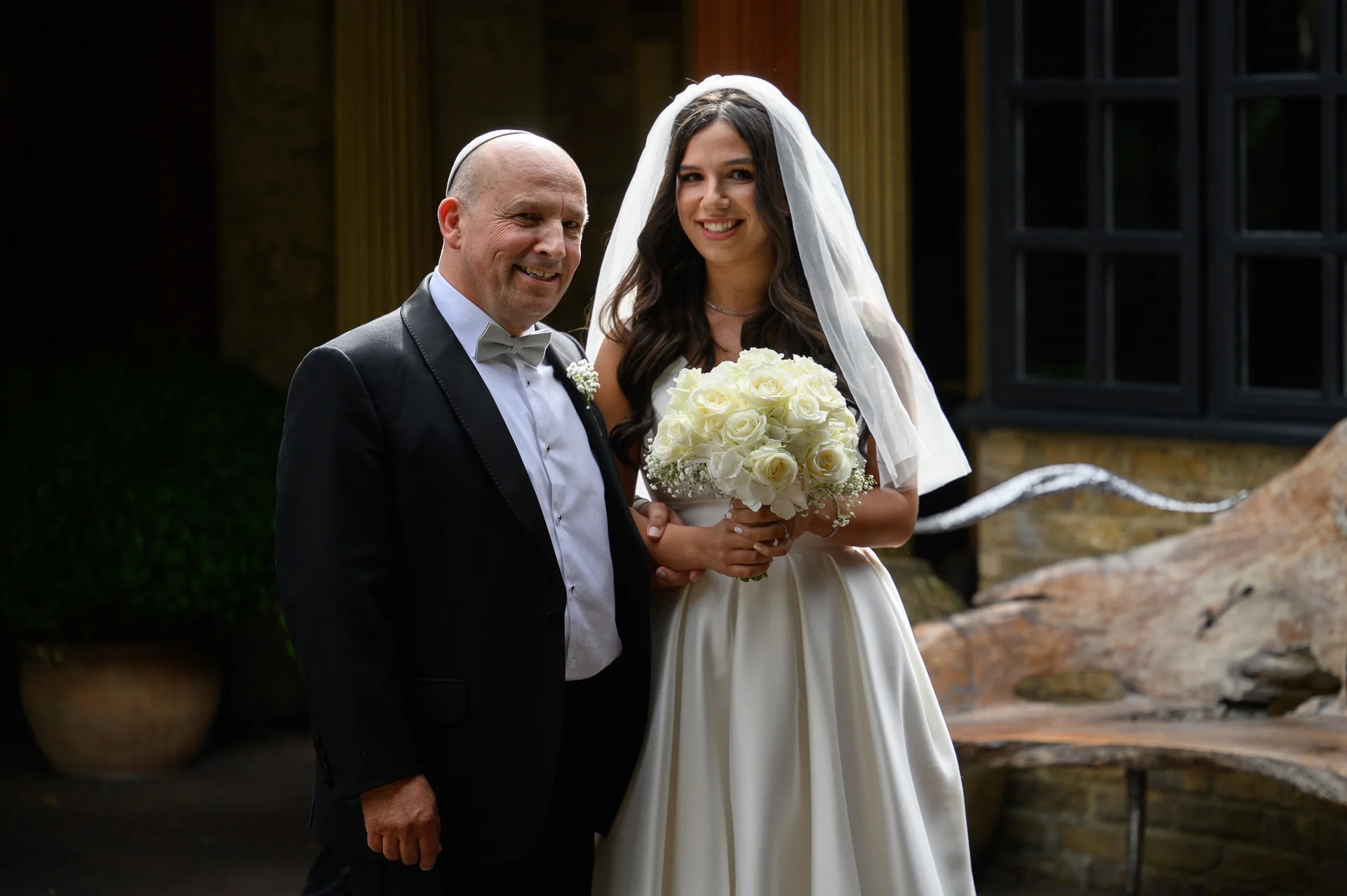 A smiling bride holding a white rose bouquet stands alongside her father wearing a black tuxedo and yarmulke.