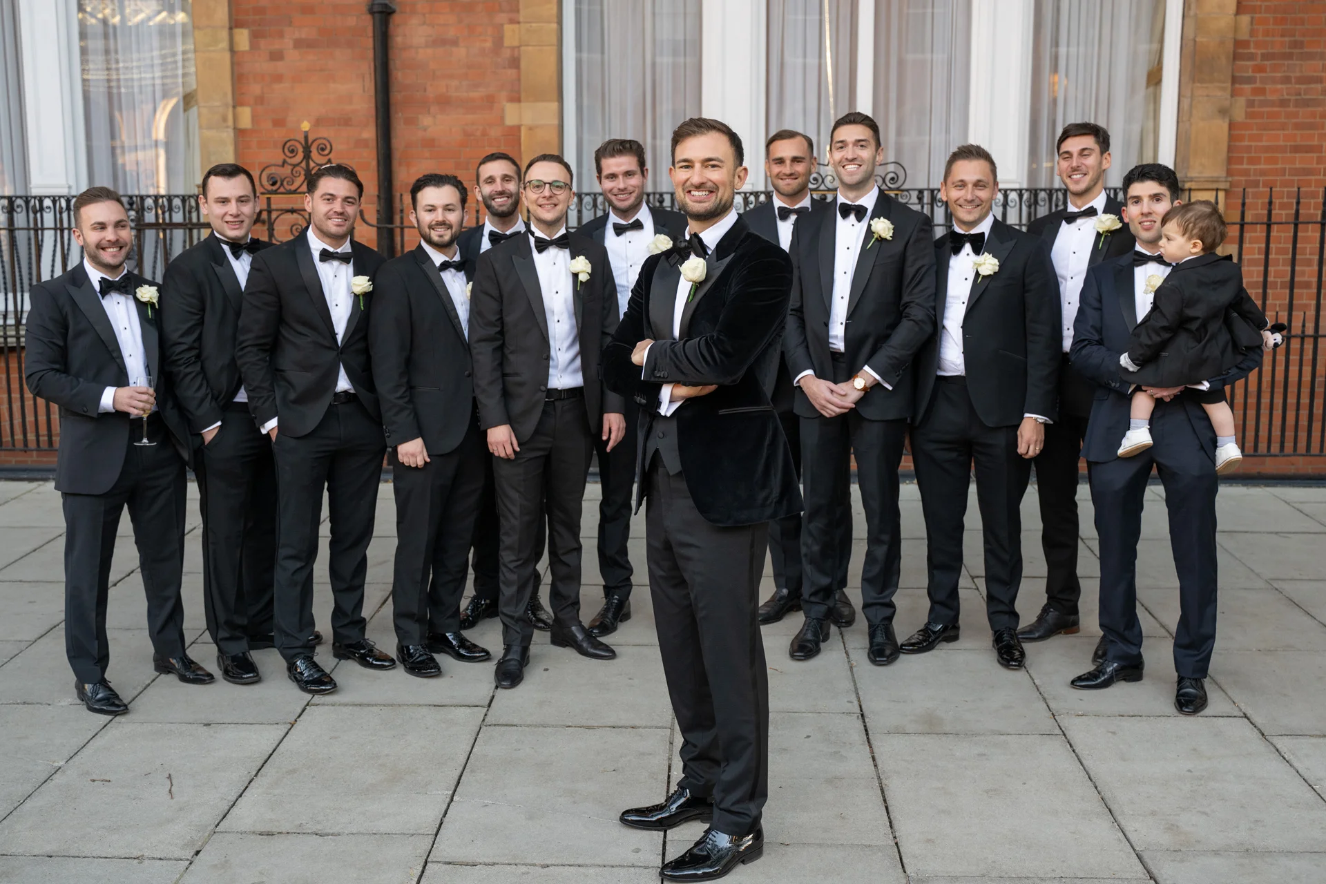 The groom stands confidently with his arms crossed in the foreground, surrounded by a large group of groomsmen wearing black tuxedos and bow ties outside a brick building.