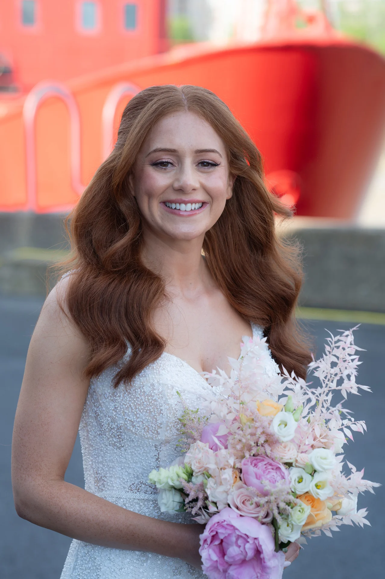A smiling red-haired bride with a half-up hairstyle and beaded wedding dress holds a pastel floral bouquet against a harbour backdrop.