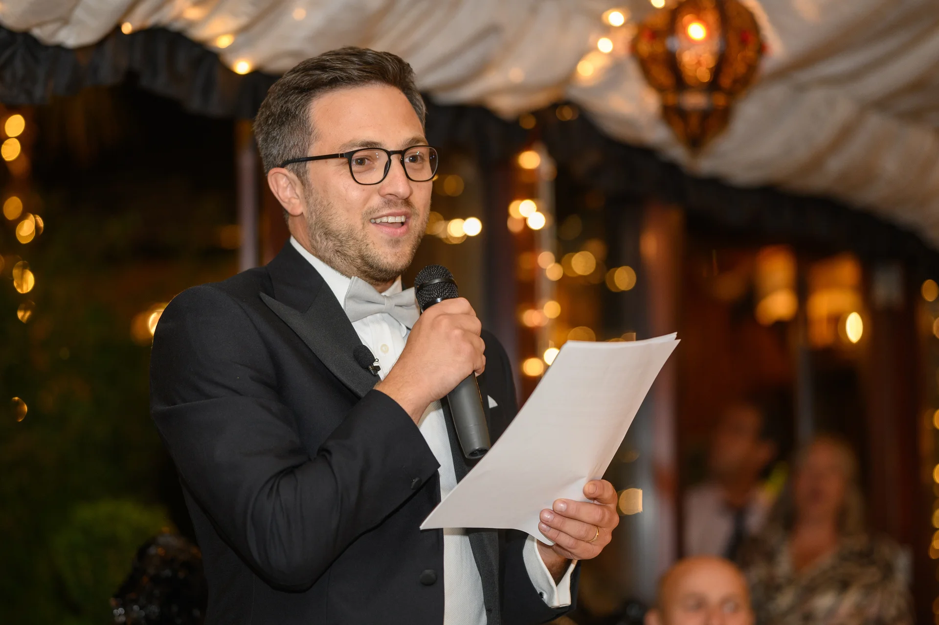 A groom in a black tuxedo and bow tie delivers his wedding speech, holding a microphone and speech notes under a canopy of warm fairy lights.