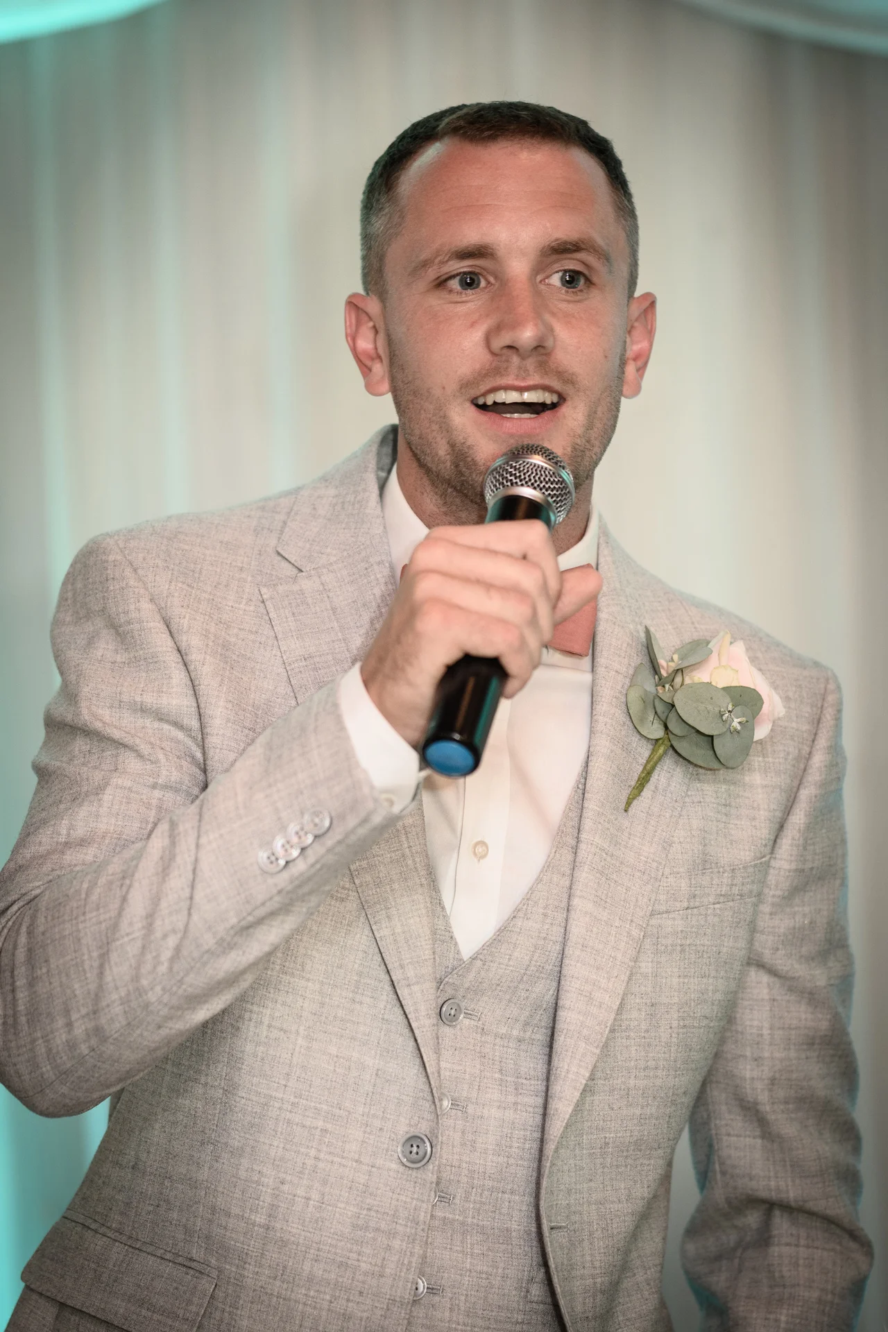 A groom wearing a light grey suit and waistcoat delivers a speech while holding a microphone at the reception.