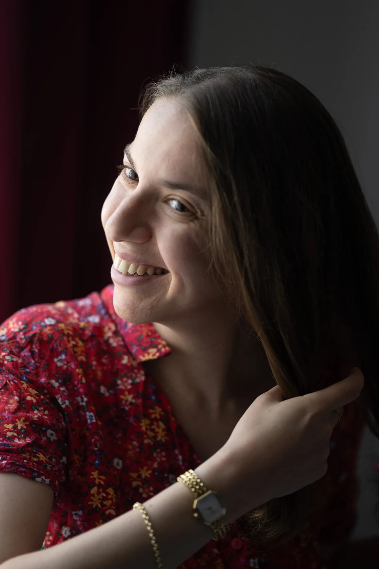 A smiling woman with long brown hair wearing a red floral dress looks over her shoulder while illuminated by soft window light.