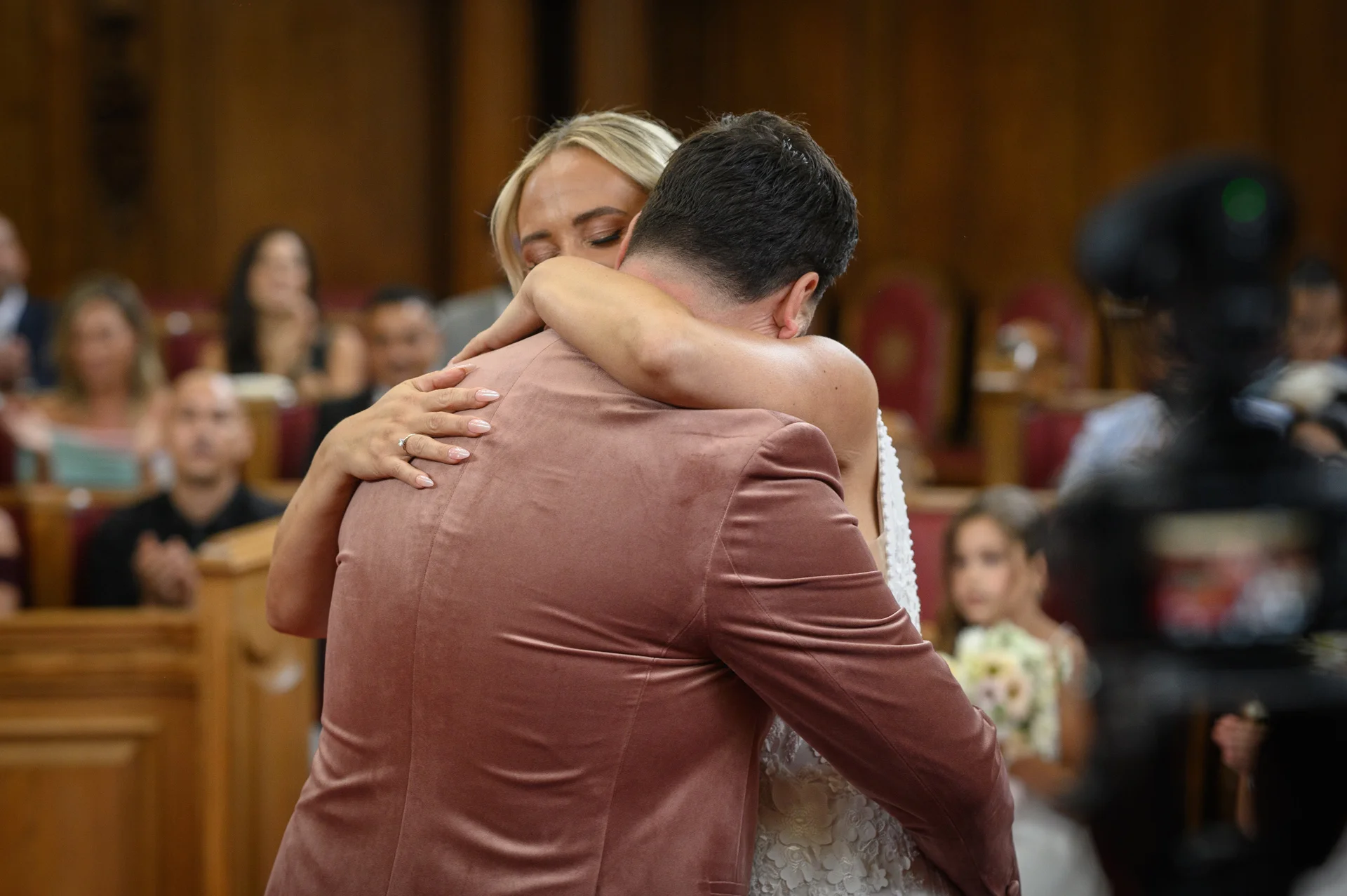 A blonde bride lovingly embraces her groom, who is wearing a stylish pink velvet suit jacket, during their emotional wedding ceremony.