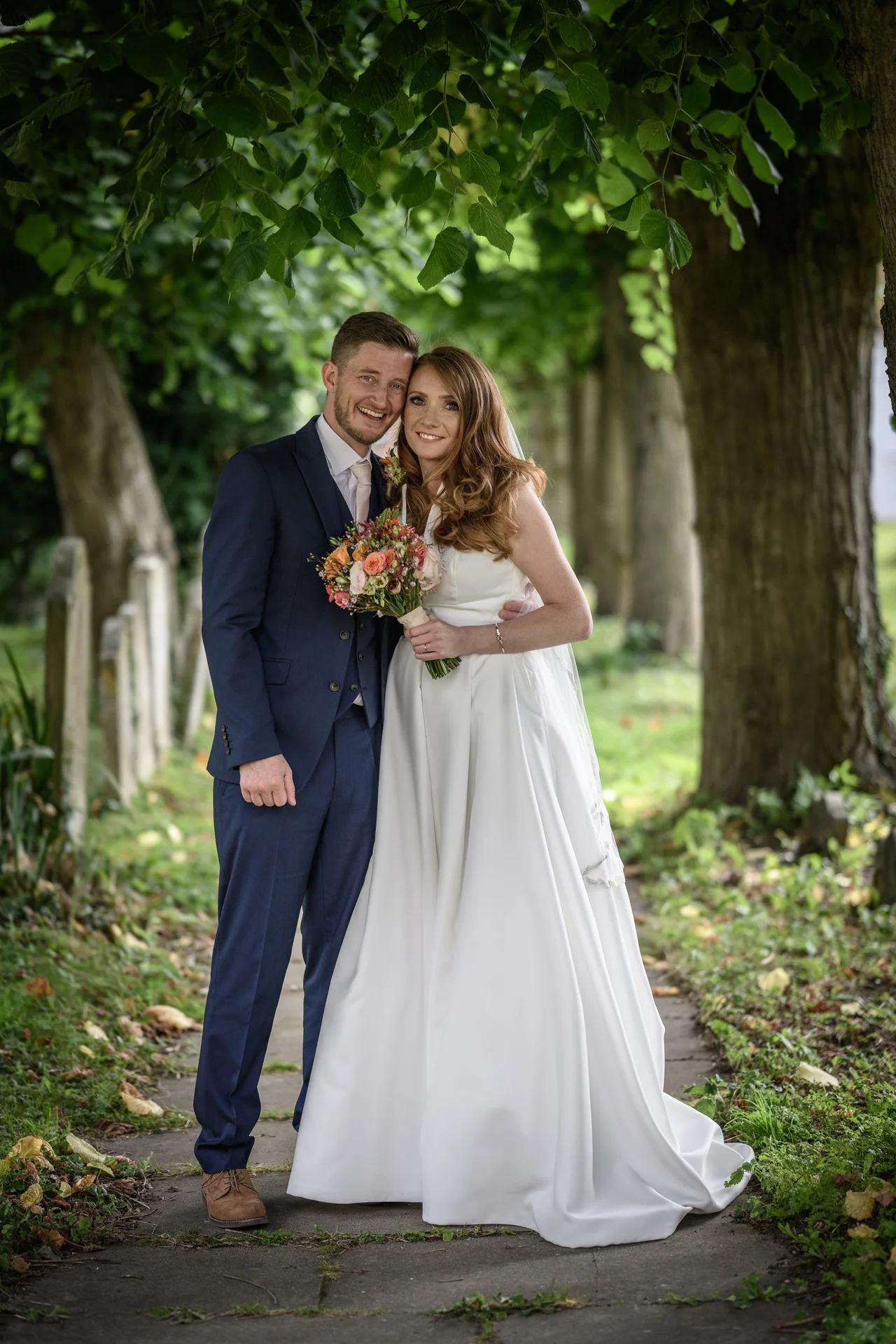 A smiling bride and groom stand together on a paved path framed by a canopy of mature trees, with the bride holding a colourful hand-tied bouquet.
