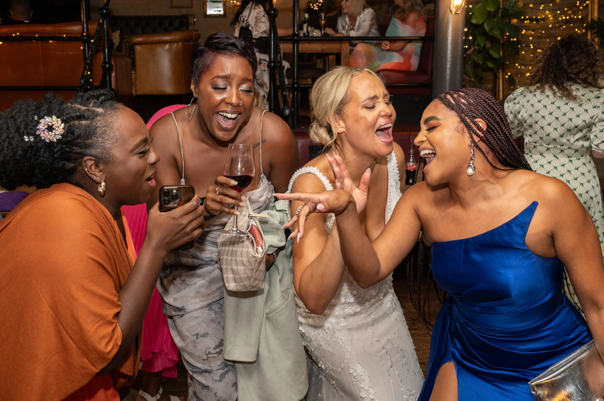 A bride and her diverse group of friends laugh uproariously together during an evening wedding reception inside a lively venue.