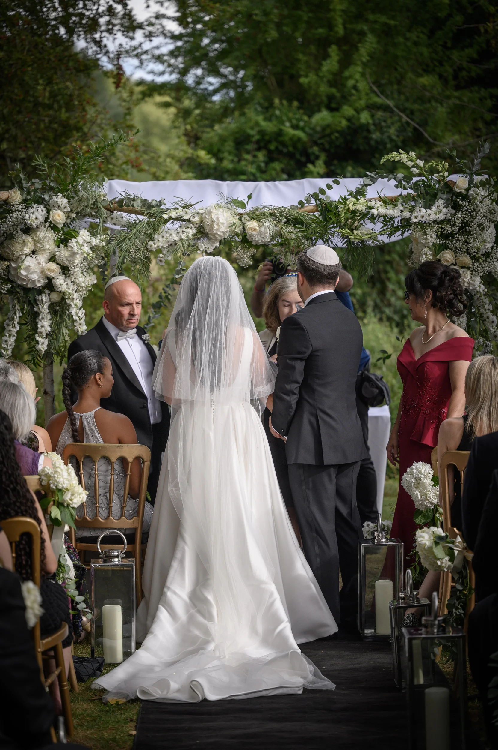 A bride and groom stand under a floral chuppah during an outdoor Jewish wedding ceremony, viewed from behind with family and guests seated along the aisle.