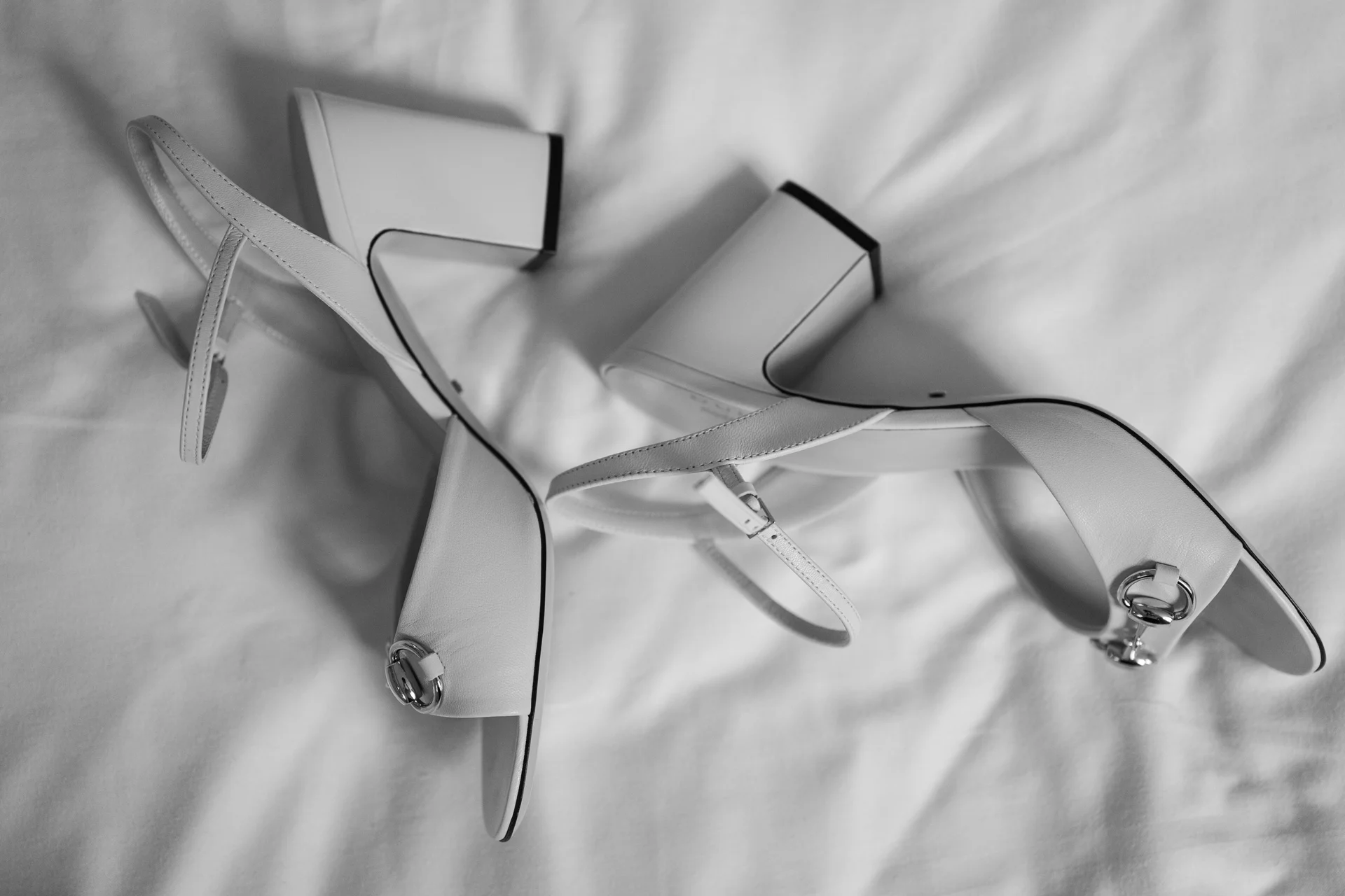 A stylish black and white flat lay capturing a pair of elegant block heel bridal sandals resting on crisp white bedding.