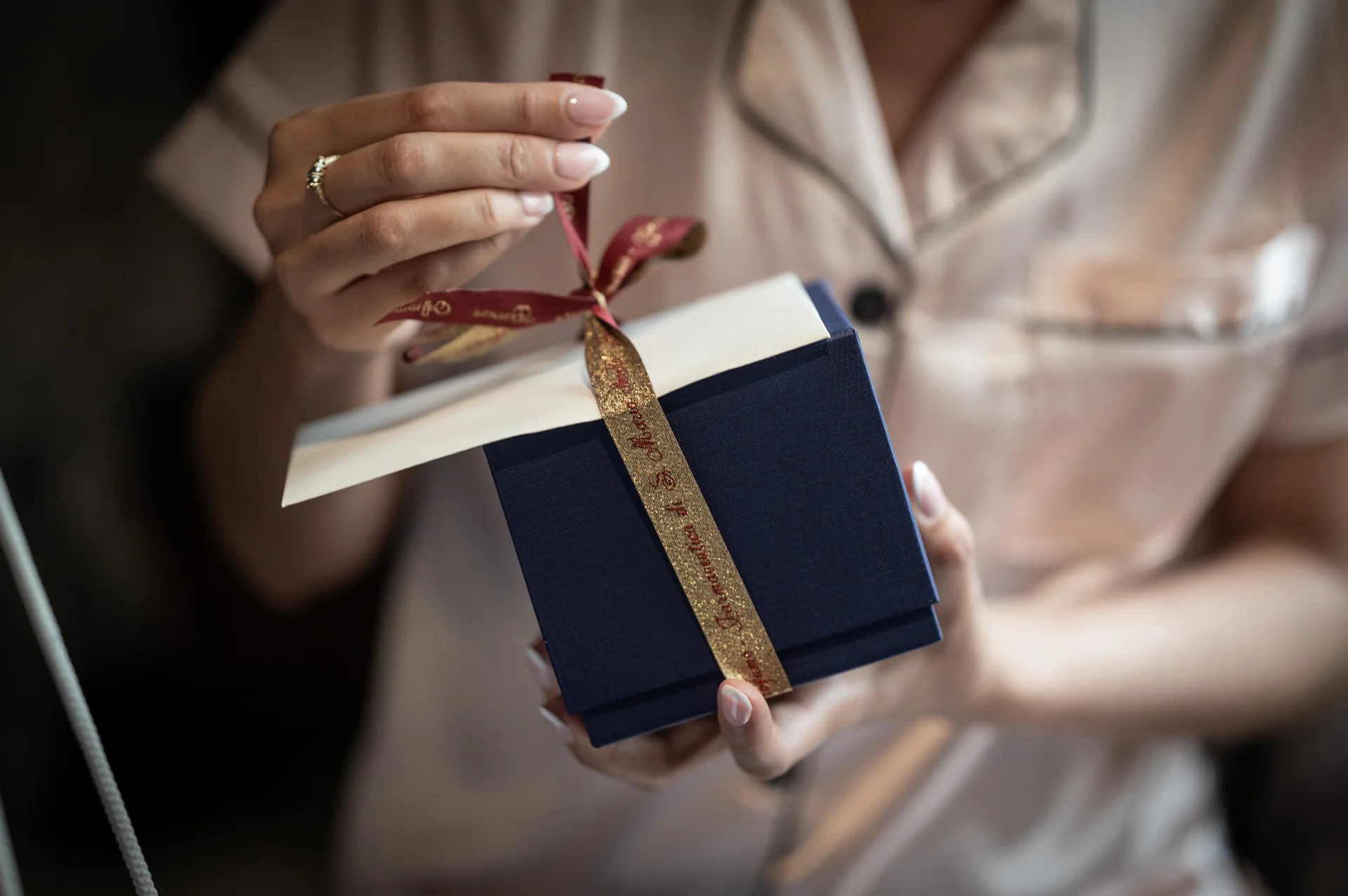 A person wearing silky pyjamas carefully unties a decorative ribbon on a small dark blue and white gift box during bridal morning preparations.