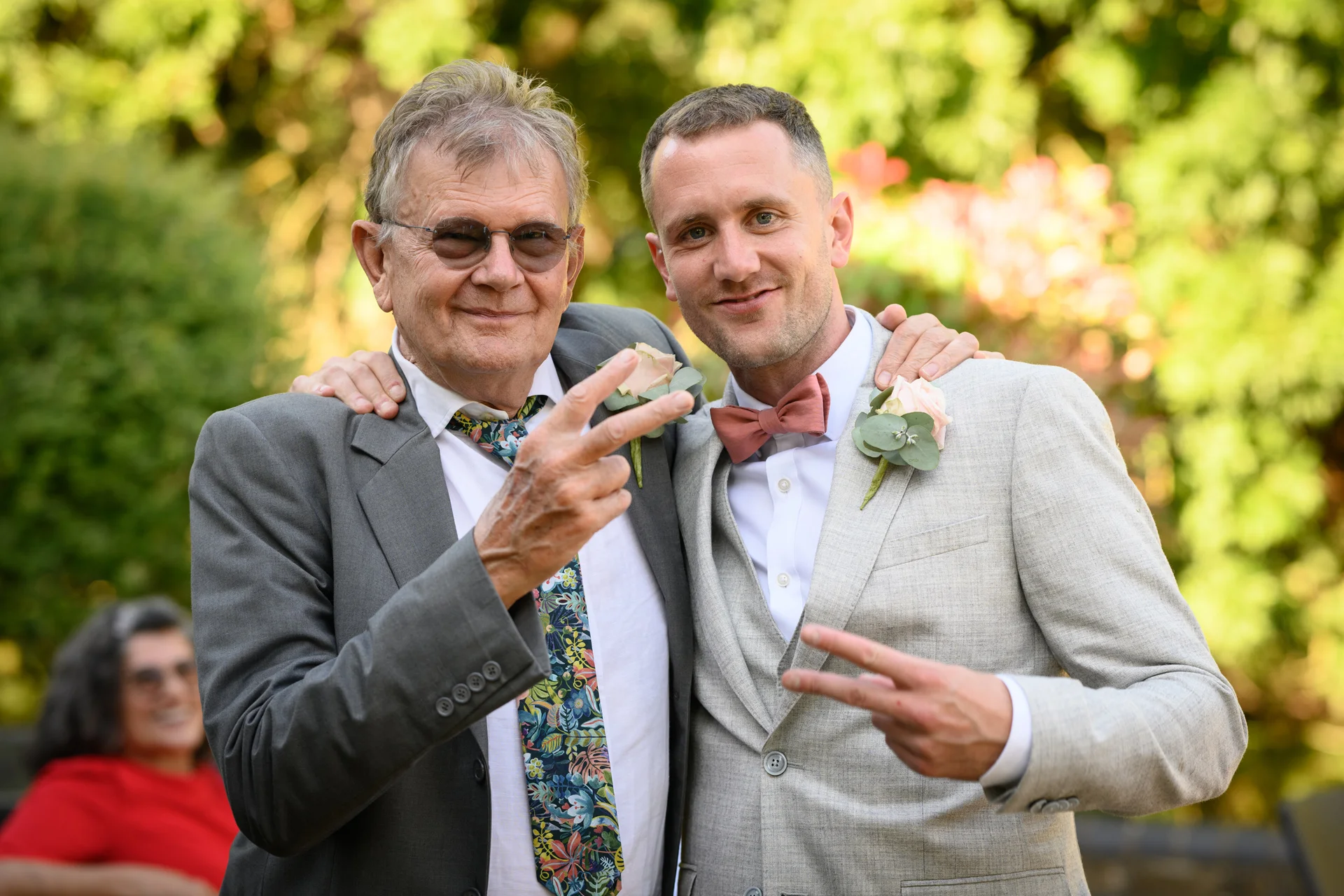 An older guest wearing sunglasses and a patterned tie poses with a groom in a light grey suit, both flashing a peace sign during the wedding celebration.