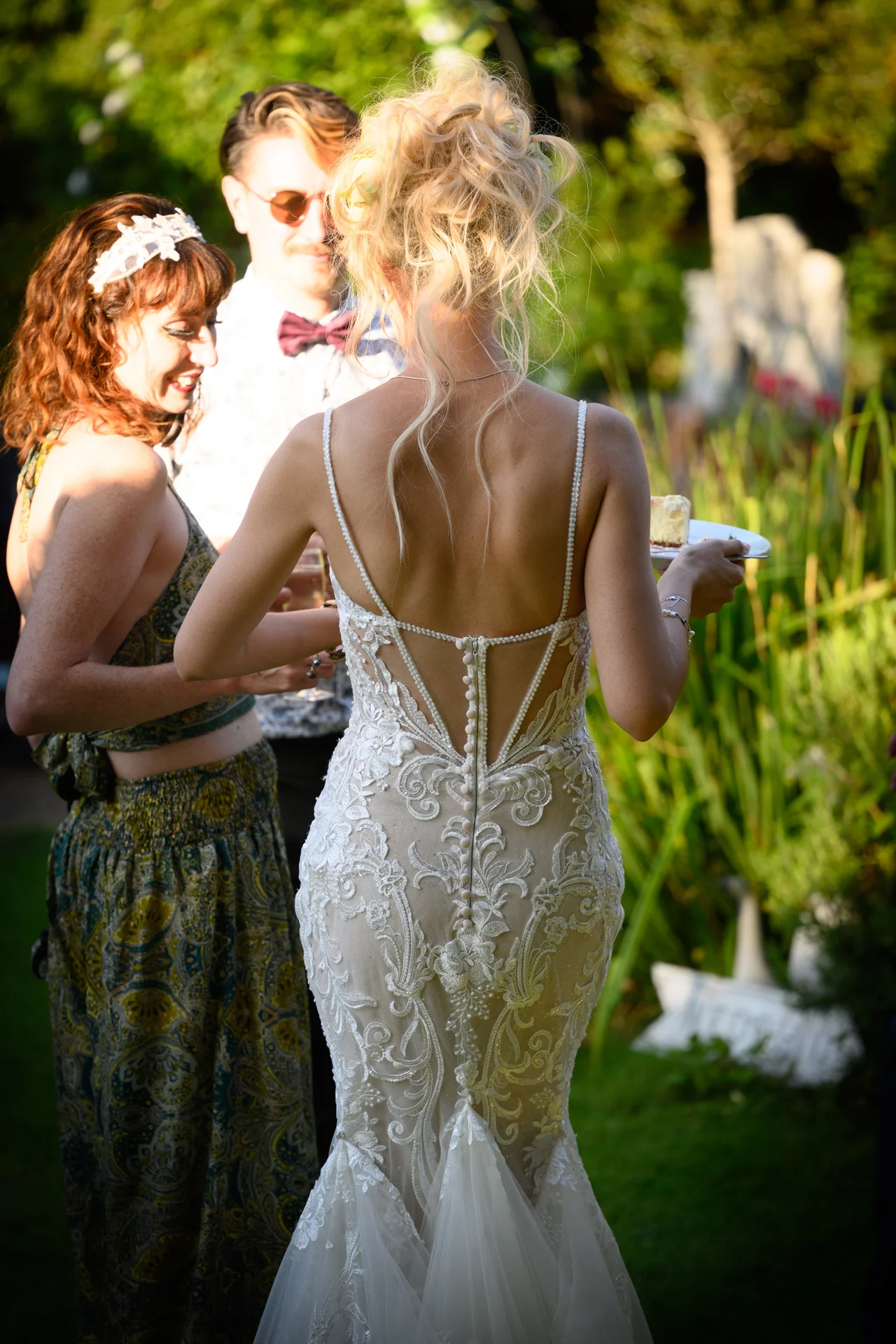 A bride in a sheer lace fit-and-flare gown with a low button back holds a plate of cake while mingling outdoors during the golden hour.