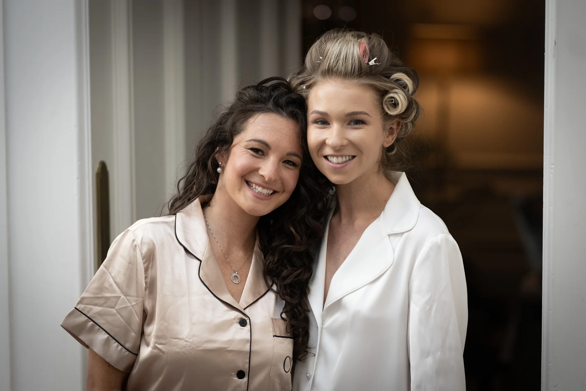 Two smiling women pose together indoors during bridal preparations, wearing satin pyjamas while one has hair rollers in her blonde locks.