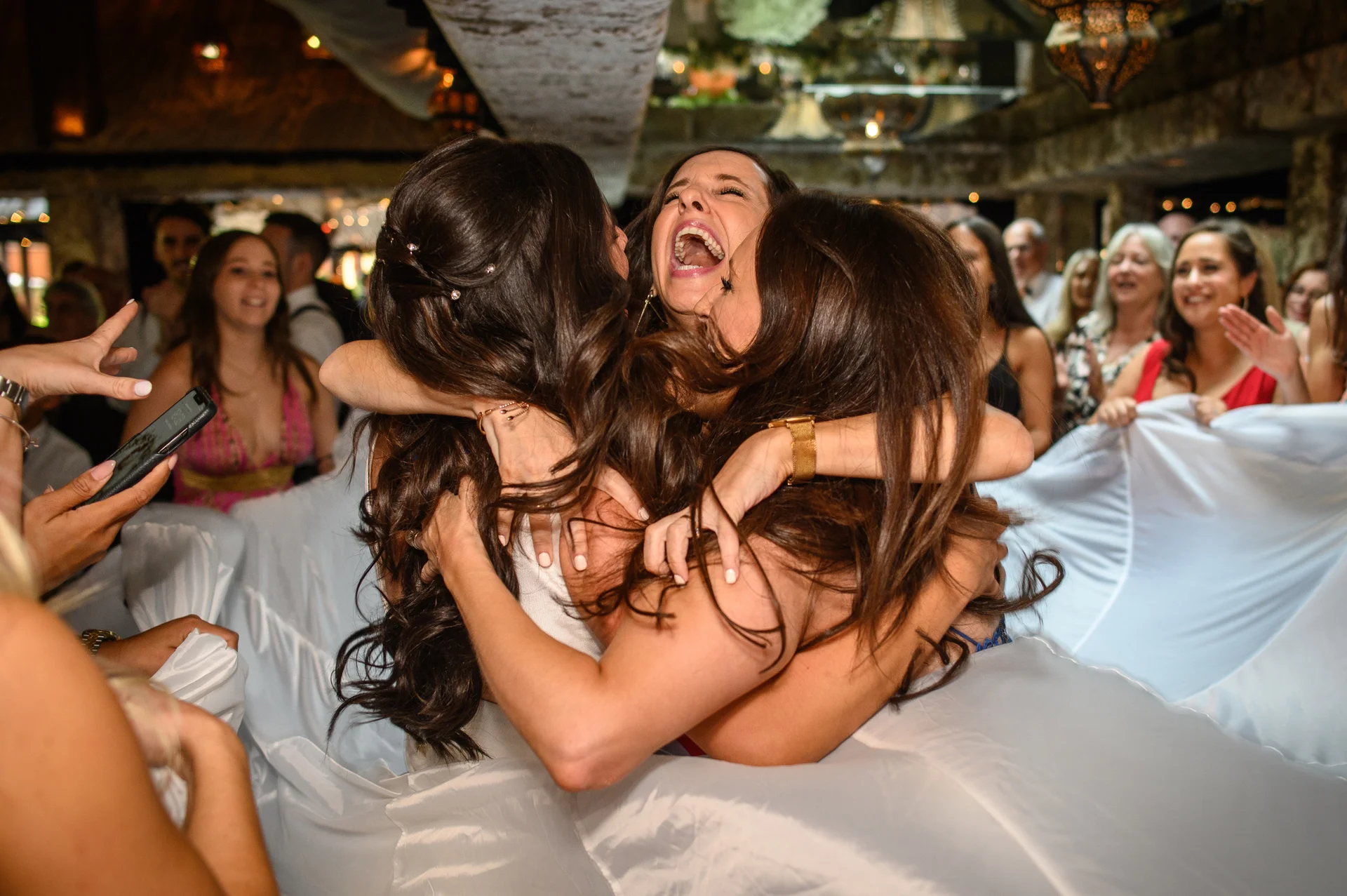 A group of joyful wedding guests laugh and celebrate together in an ecstatic embrace while lifted high in the air during the reception.