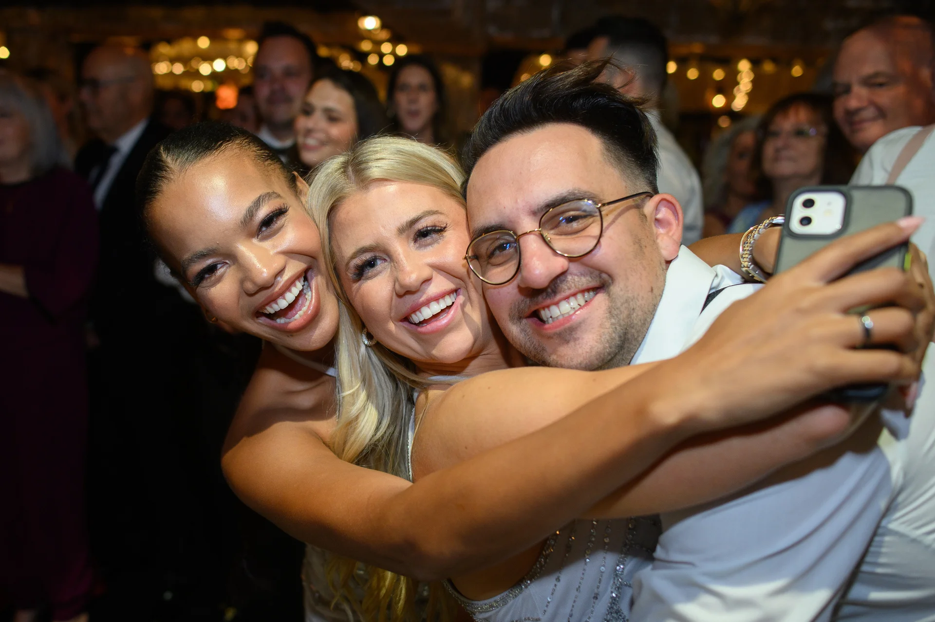 Three wedding guests enthusiastically embrace and smile together while taking a selfie on a smartphone at a lively evening reception.