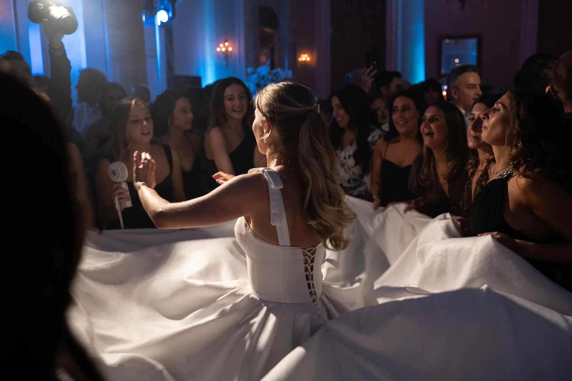 A bride in a white ball gown with a lace-up back stands on the dance floor surrounded by cheering guests holding out her dress.