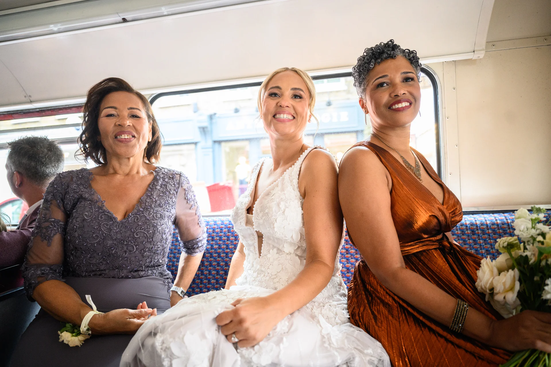 A joyful bride in a floral applique gown sits back to back with a bridesmaid holding a bouquet aboard a vintage London bus, accompanied by another guest.