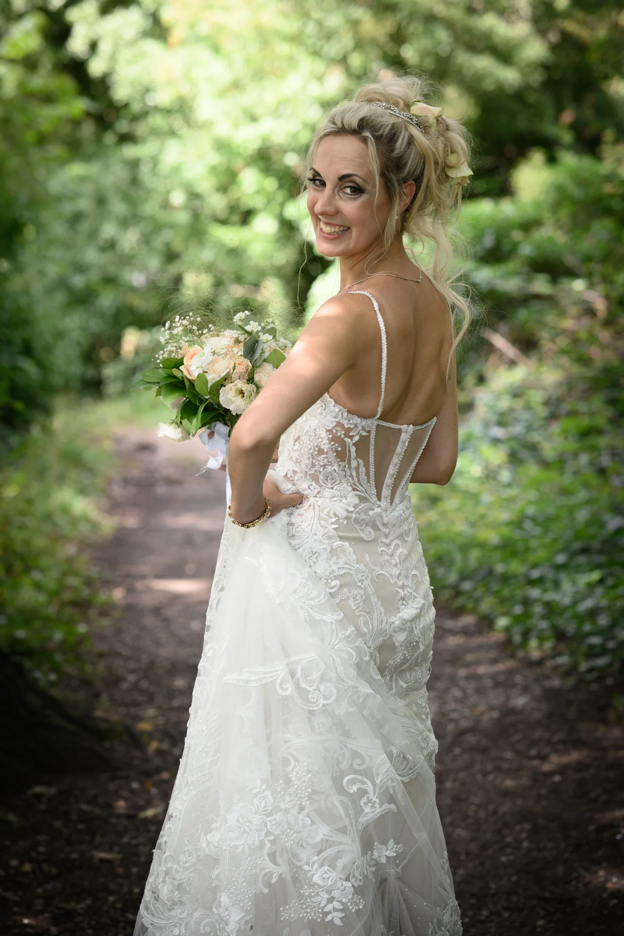 A smiling bride wearing an intricate lace gown with a sheer corset back looks over her shoulder while holding a hand-tied bouquet in a lush woodland setting.