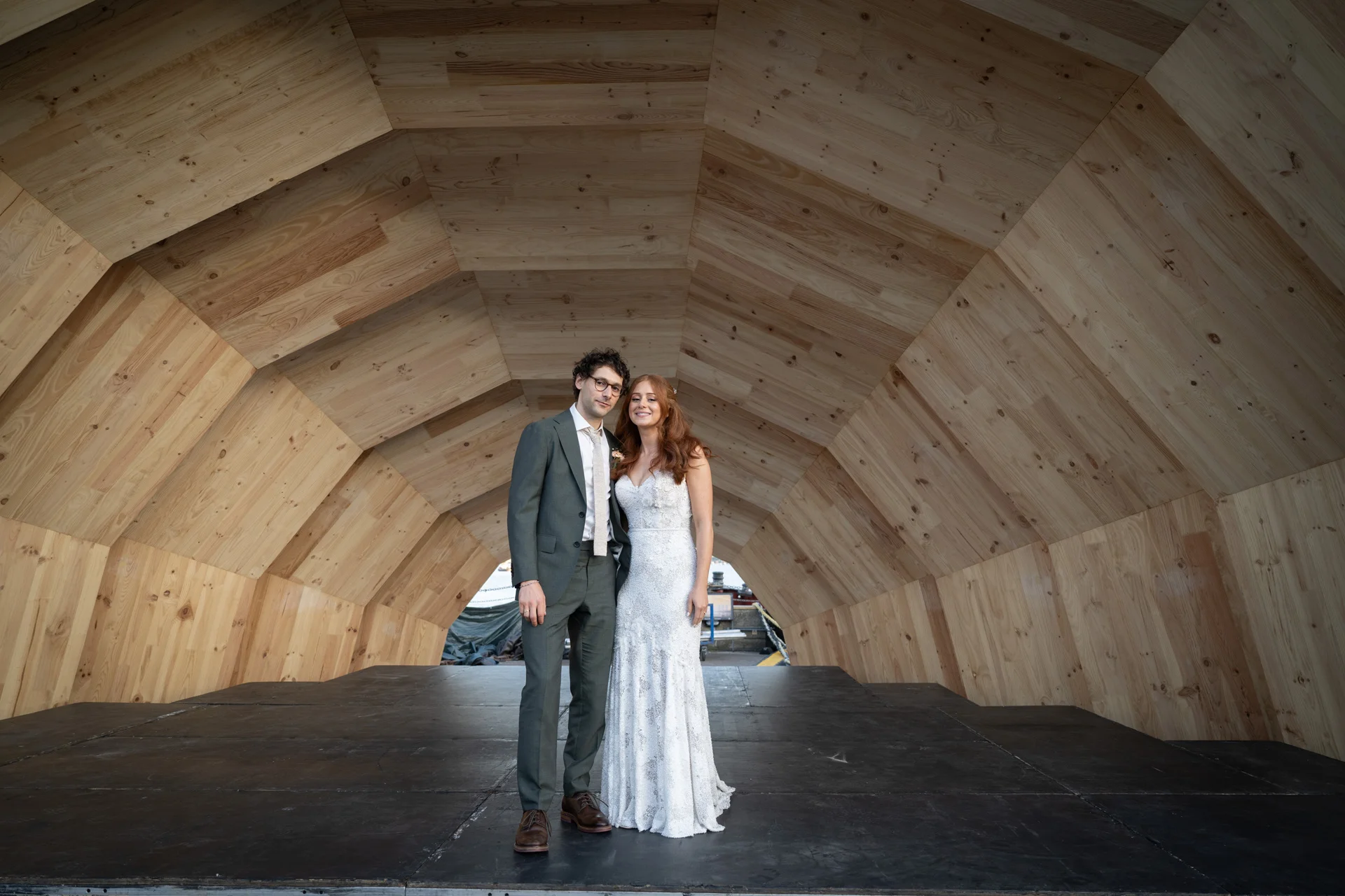 A bride and groom pose inside a striking wooden geometric tunnel structure at their modern wedding venue.