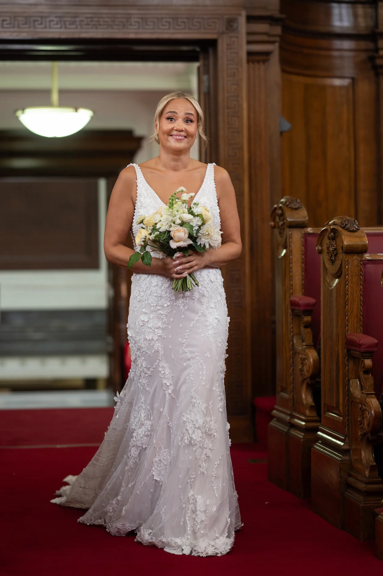 A smiling bride holding a floral bouquet walks down the red-carpeted aisle of a wooden church during her ceremony.