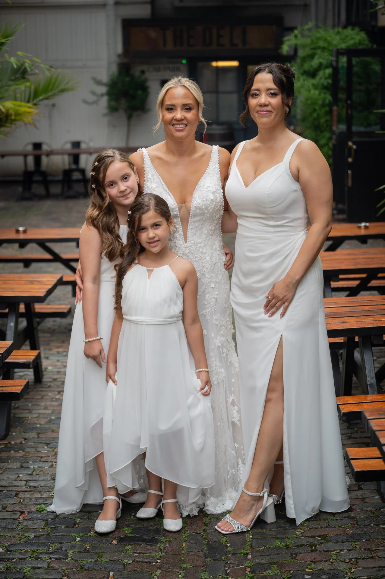 Two brides pose together outdoors in a cobbled courtyard alongside two young flower girls wearing matching white dresses.