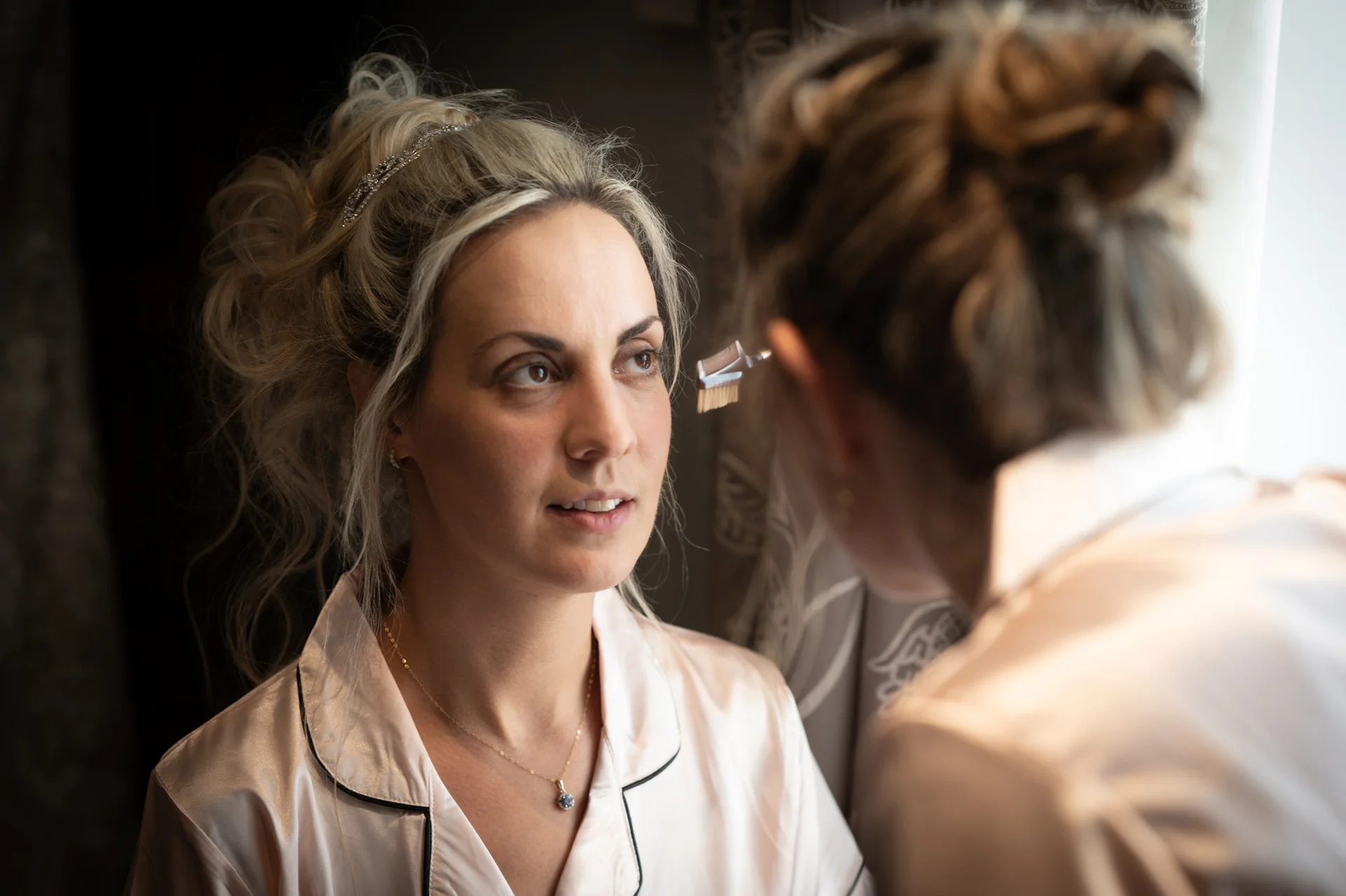 A blonde bride having her makeup applied by a stylist while wearing pyjamas and a delicate pendant necklace during morning preparations.