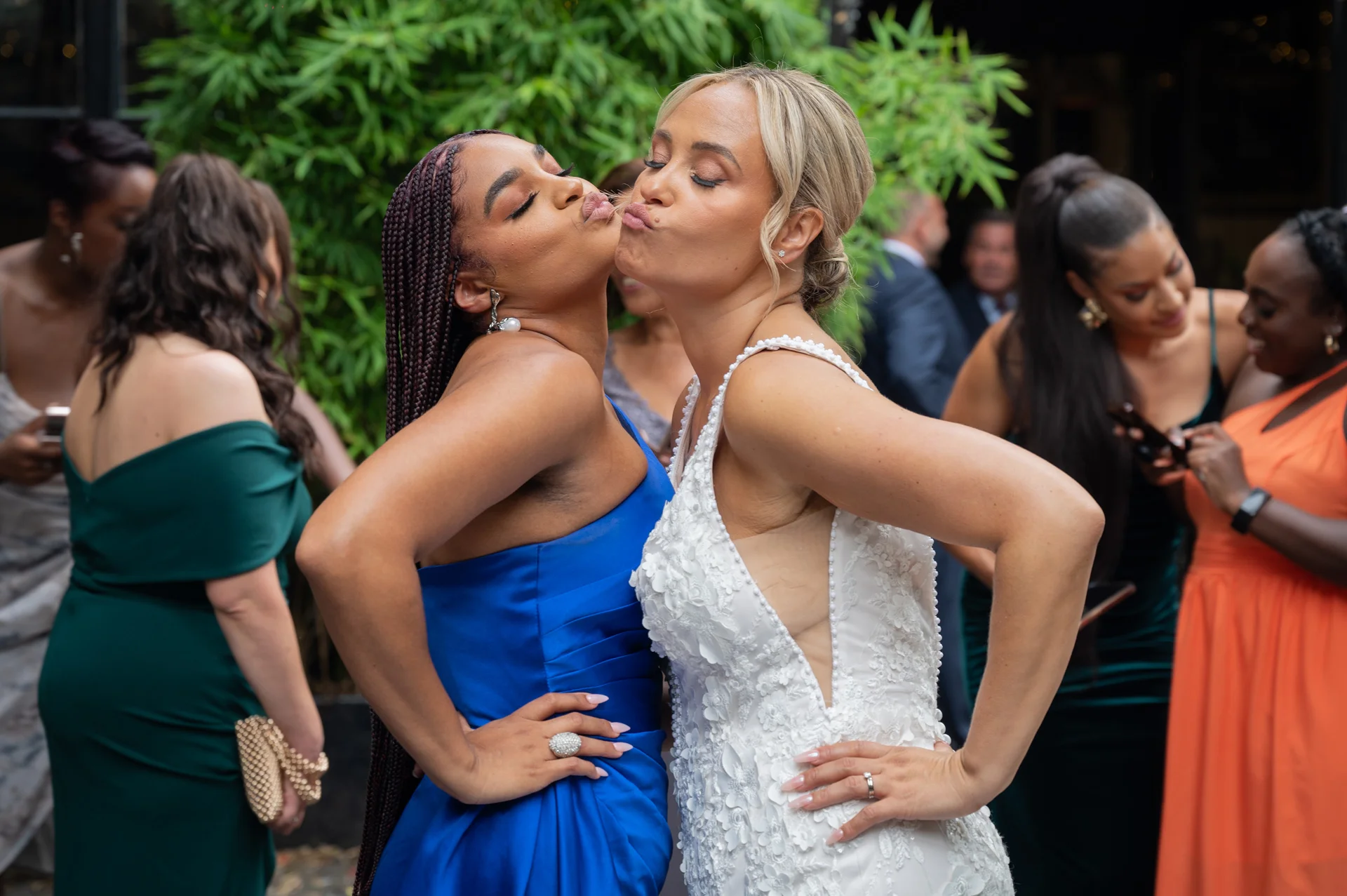 Two brides in stylish wedding attire share a playful, joyful kiss with pursed lips while standing outdoors amidst wedding guests.