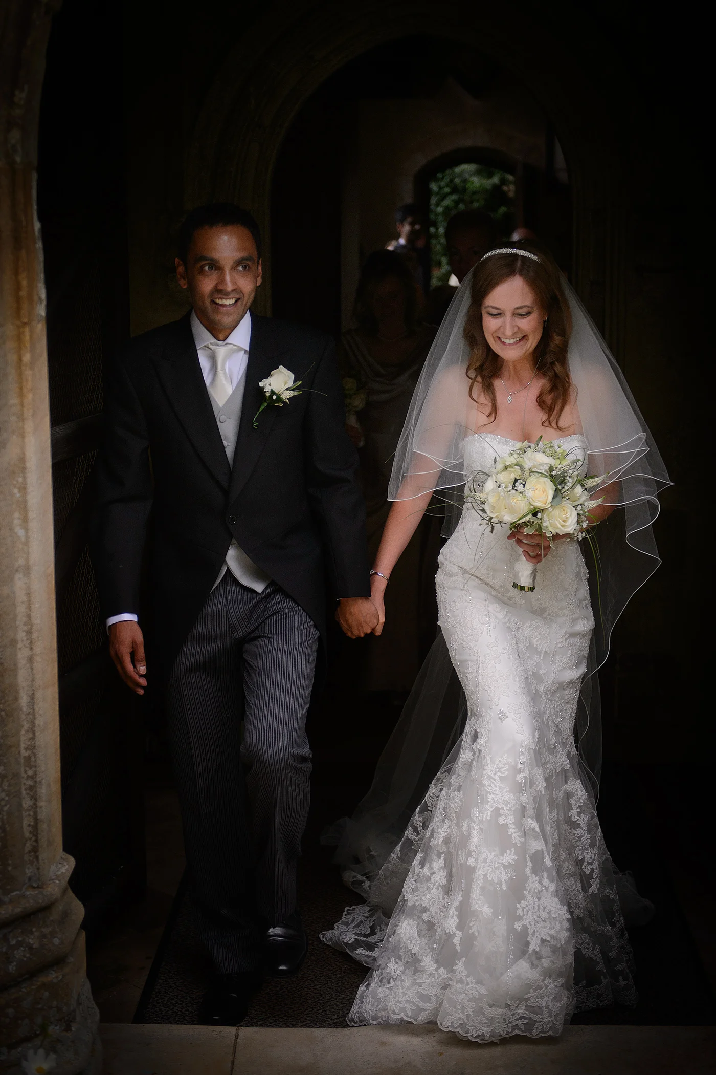 A smiling bride and groom walk hand in hand through a stone church archway, with the bride wearing a lace fit and flare gown and a veil.