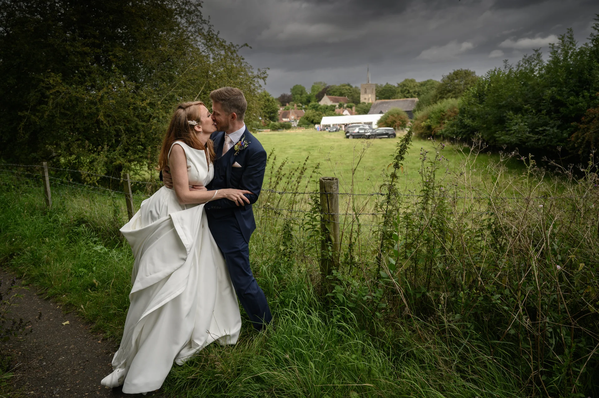 Newlyweds share a romantic kiss by a rural fence line with a dramatic stormy sky and countryside landscape in the background.