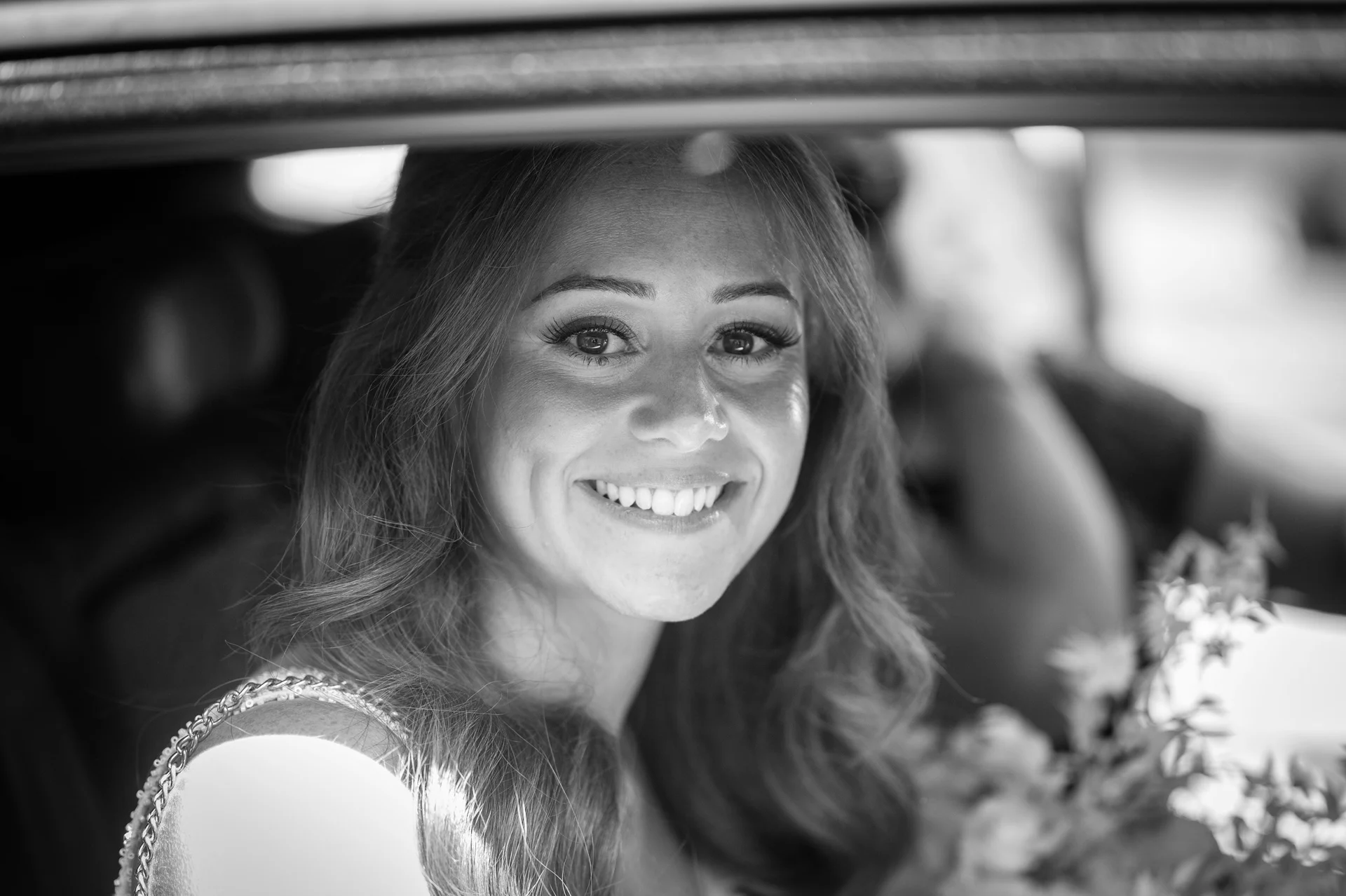 A black and white portrait of a smiling bride looking through the car window on her wedding day.
