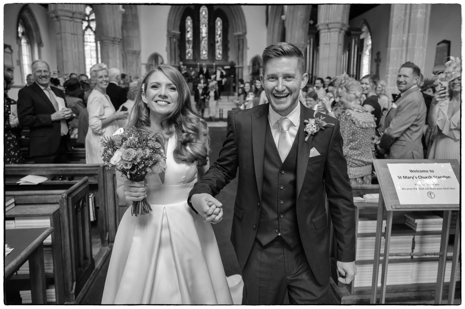 A joy-filled black and white photograph of a bride and groom walking down the aisle together after their church ceremony, smiling broadly while holding hands.