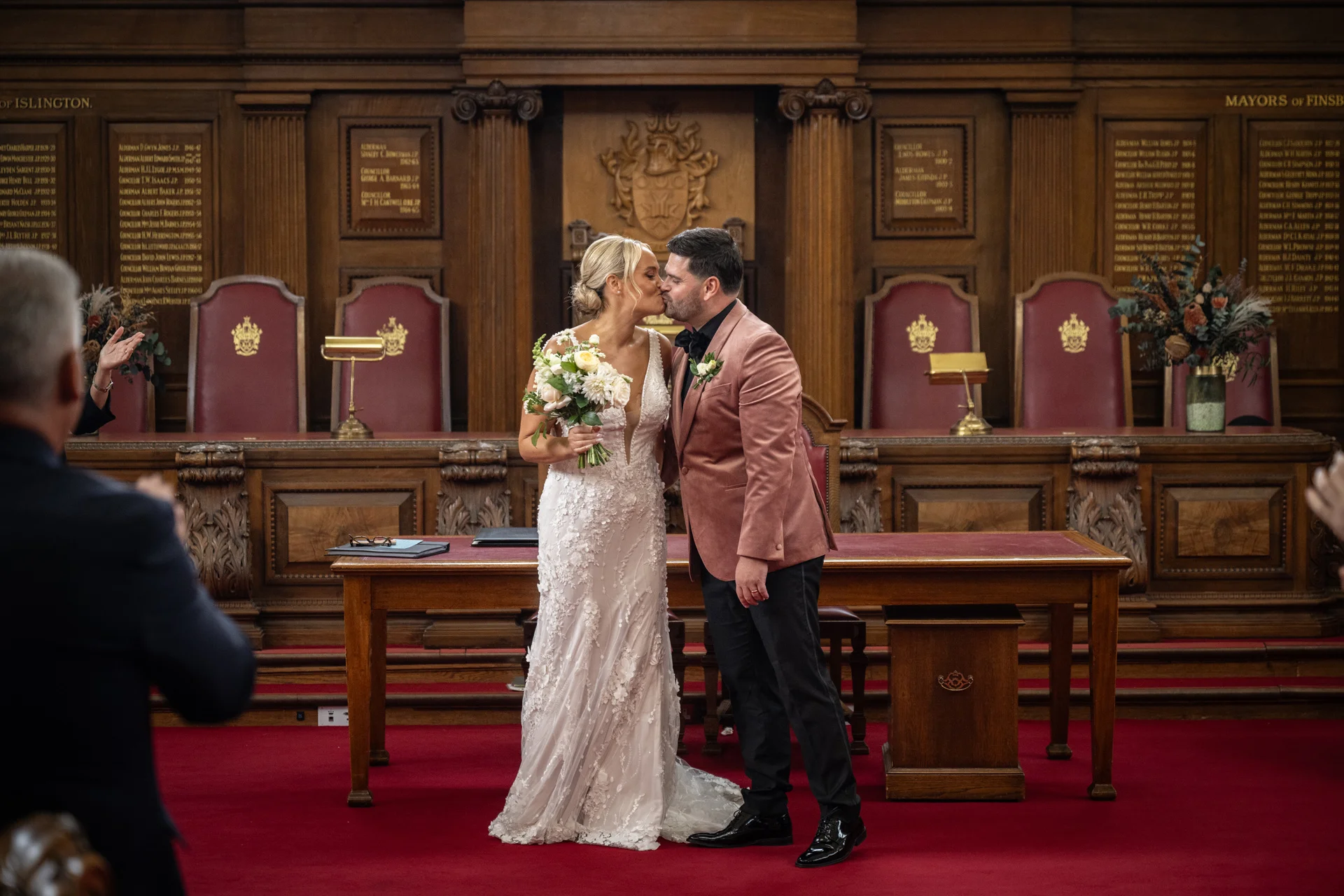A newlywed couple share their first kiss in a wood-panelled council chamber, with the bride holding a bouquet of white flowers and wearing a lace gown alongside the groom in a pink velvet jacket.