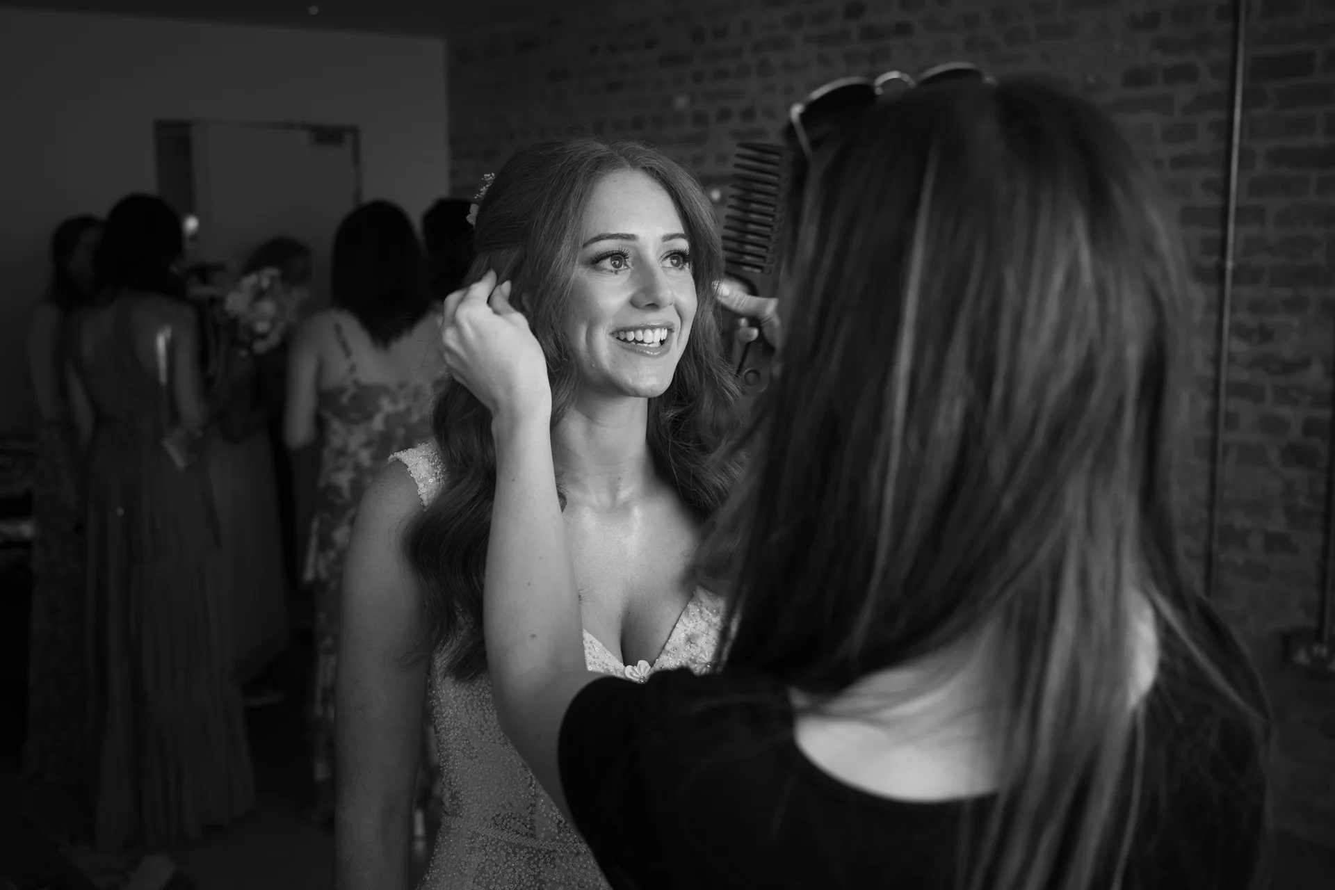 A smiling bride receives final hair styling touches from a stylist while bridesmaids gather in the background during morning preparations.